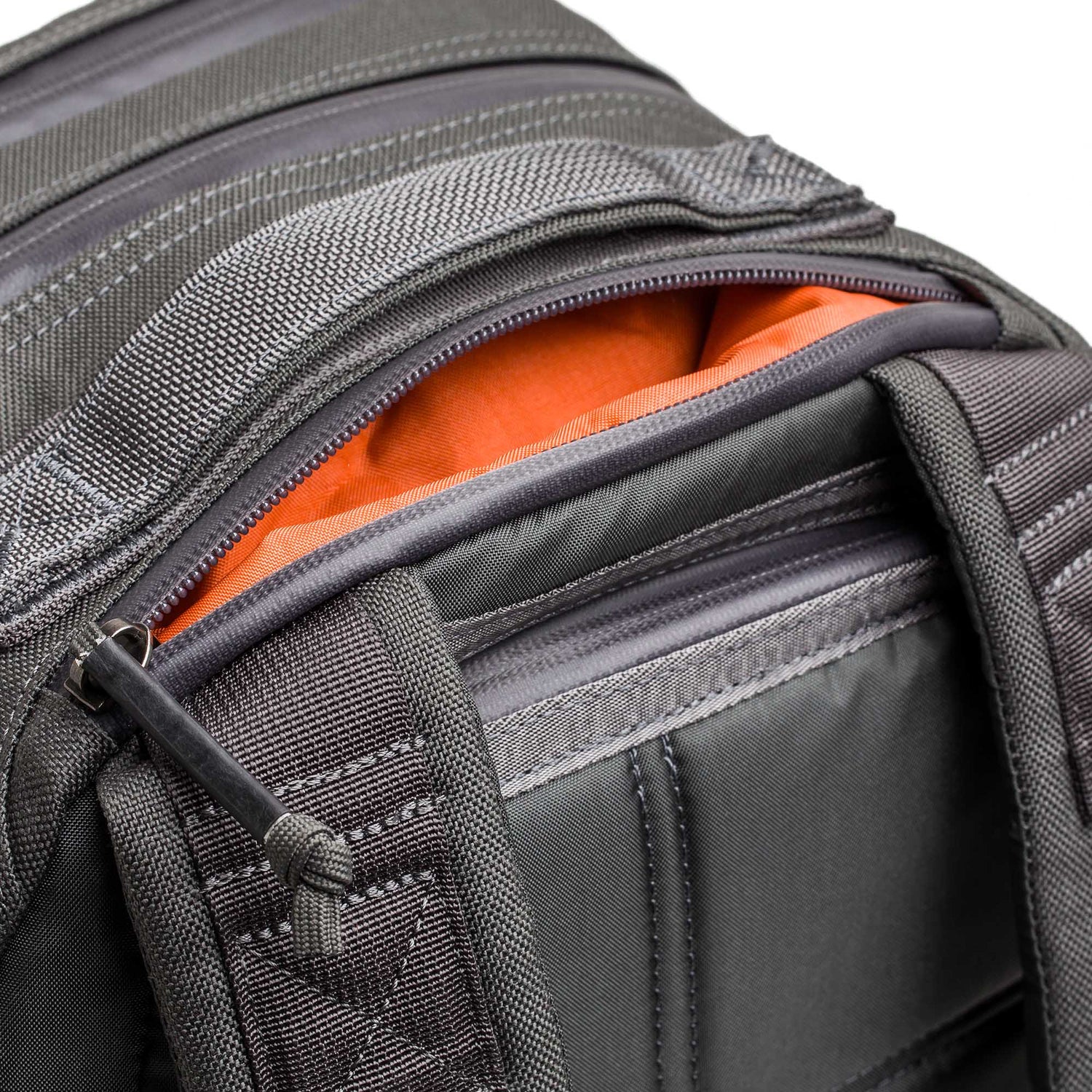 A close-up of the Double Compartment Bullet Ruck - Ballistic Nylon Cordura - 18L in gray shows an orange item in a partially open zippered pocket and highlights its bombproof laptop compartment.