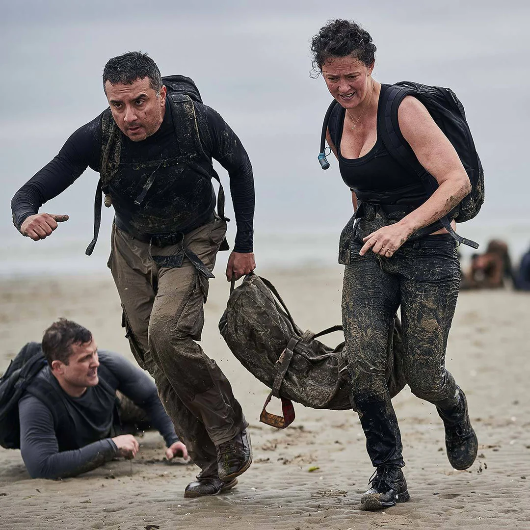 Two people in muddy clothes run on a beach, dragging a heavy bag; another person lies on the sand.