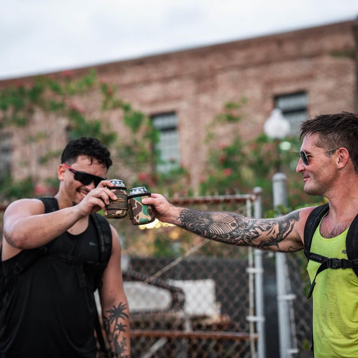 Two men in GORUCK gear cheers with camo can covers outdoors, urban backdrop