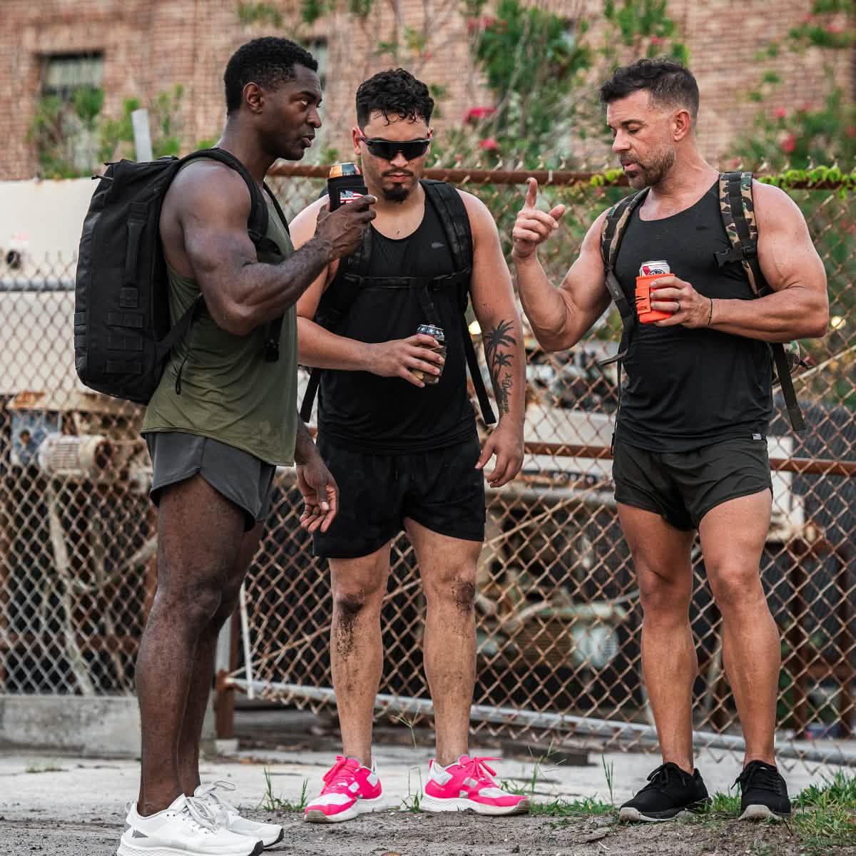 Three men in GORUCK athletic gear and backpacks stand outdoors, holding cans with beer jackets.