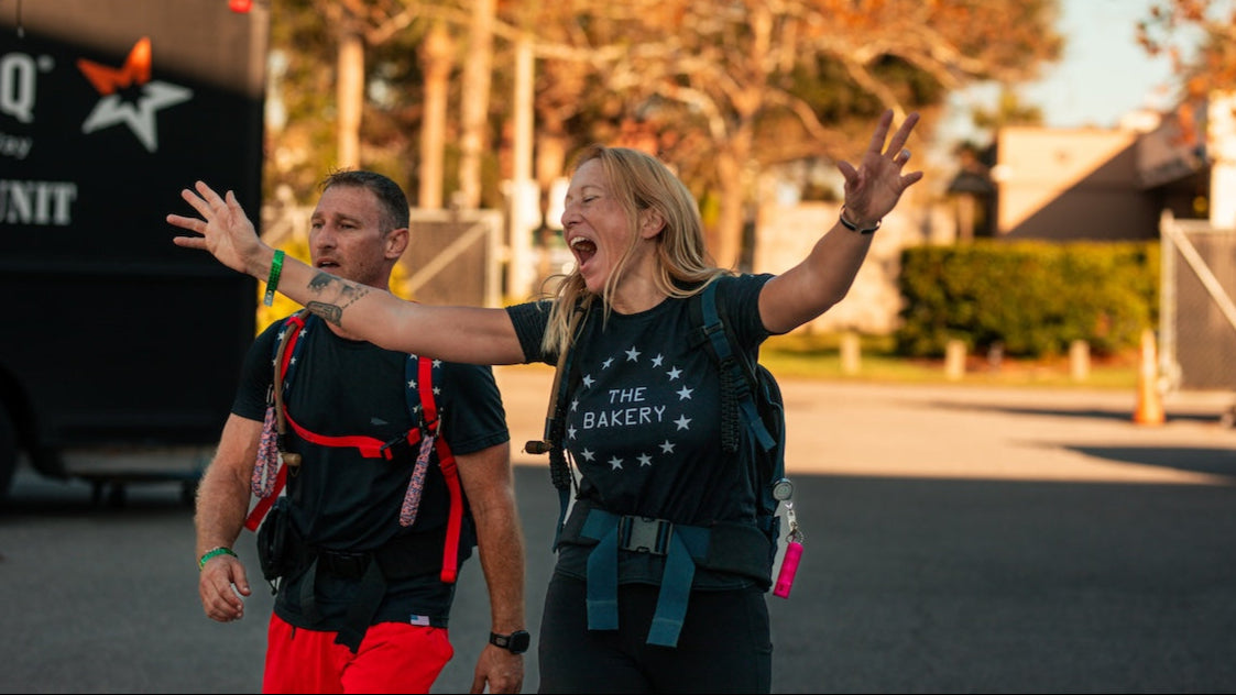 A woman with arms raised and smiling, next to a man, both wearing backpacks outdoors.