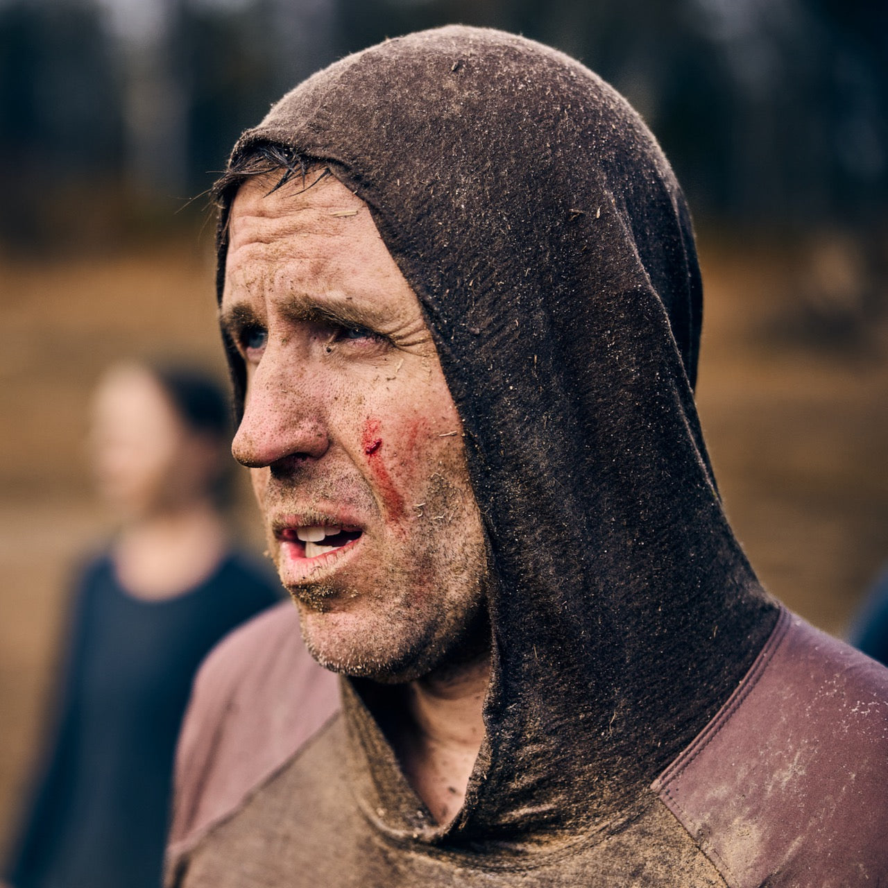 A concerned man with a scraped face stands outdoors in a muddy Men’s Commando Pullover - Merino Wool, with blurred people in the background.