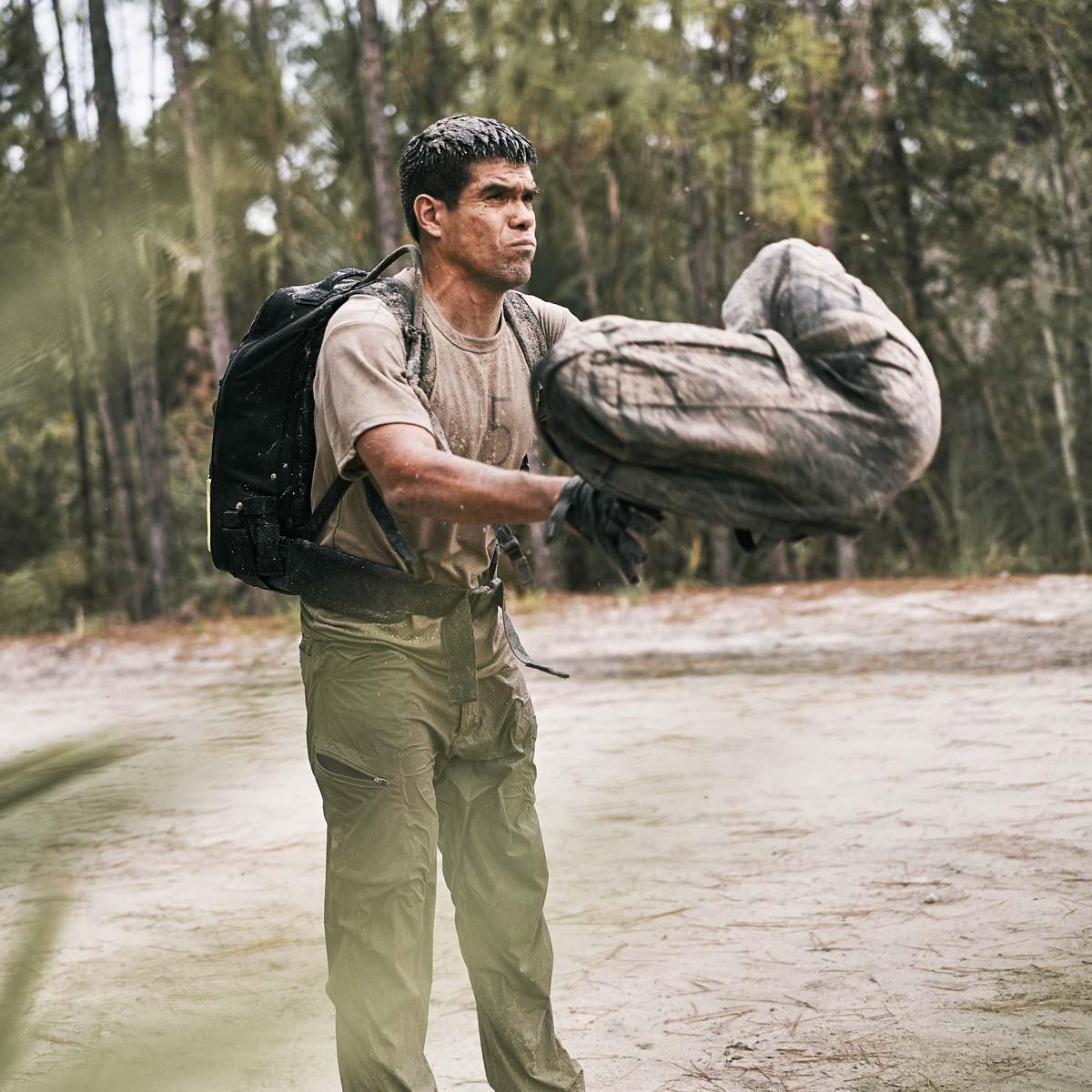 Man in GORUCK pants and backpack tossing a sandbag outdoors during rucking workout