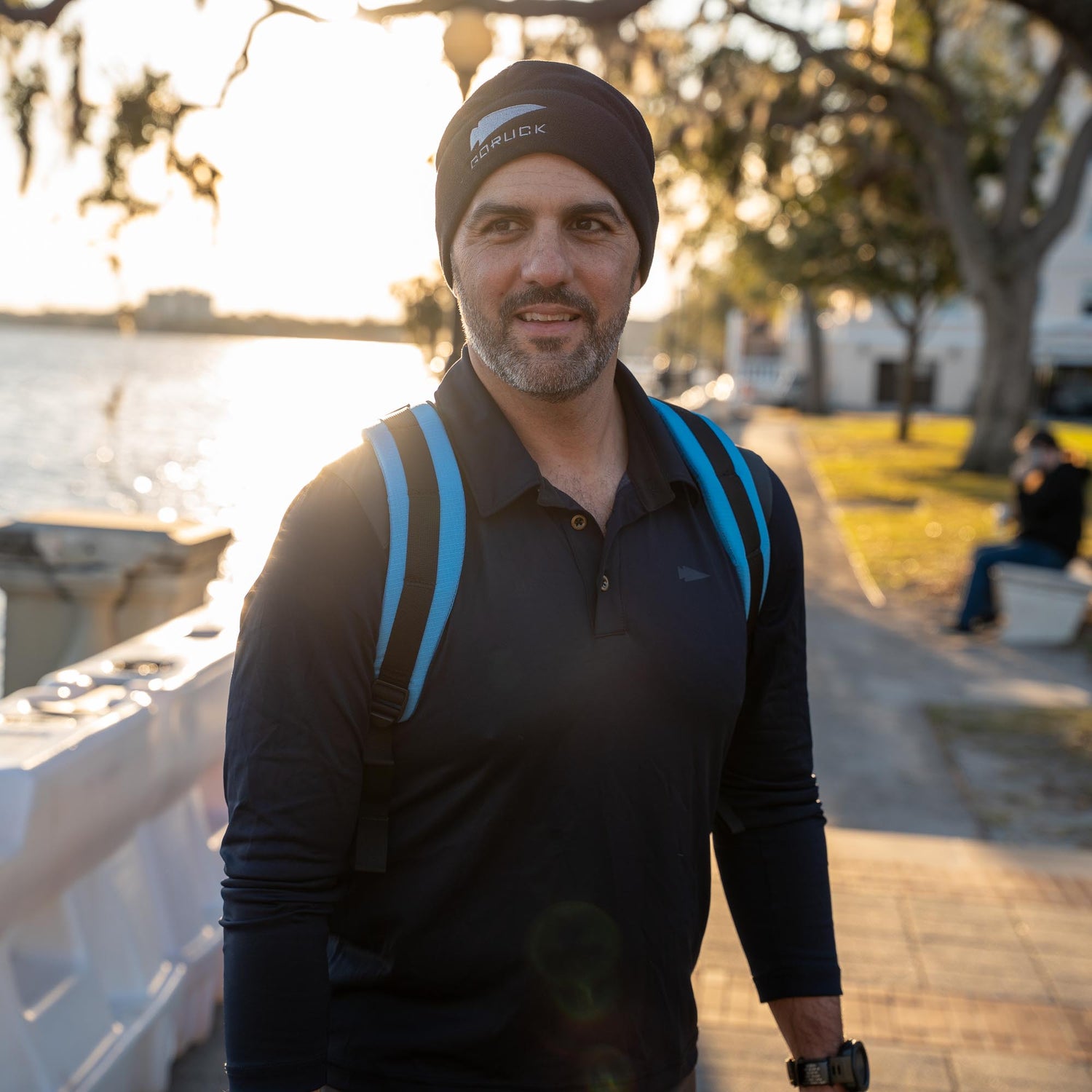 A man wearing a Men's Long Sleeve Rucking Polo and beanie with a blue backpack walks by the waterfront at sunset.