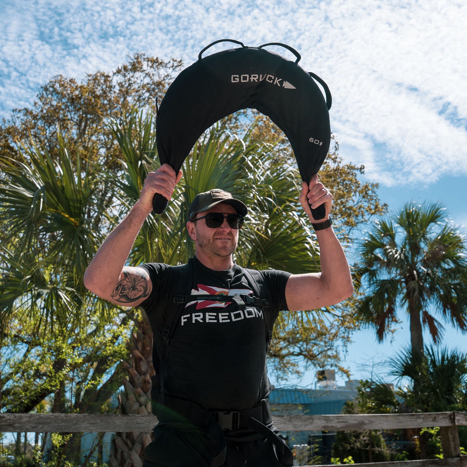A man outdoors lifts a Bulgarian Sandbag with reinforced handles overhead, wearing sunglasses and a shirt that reads
