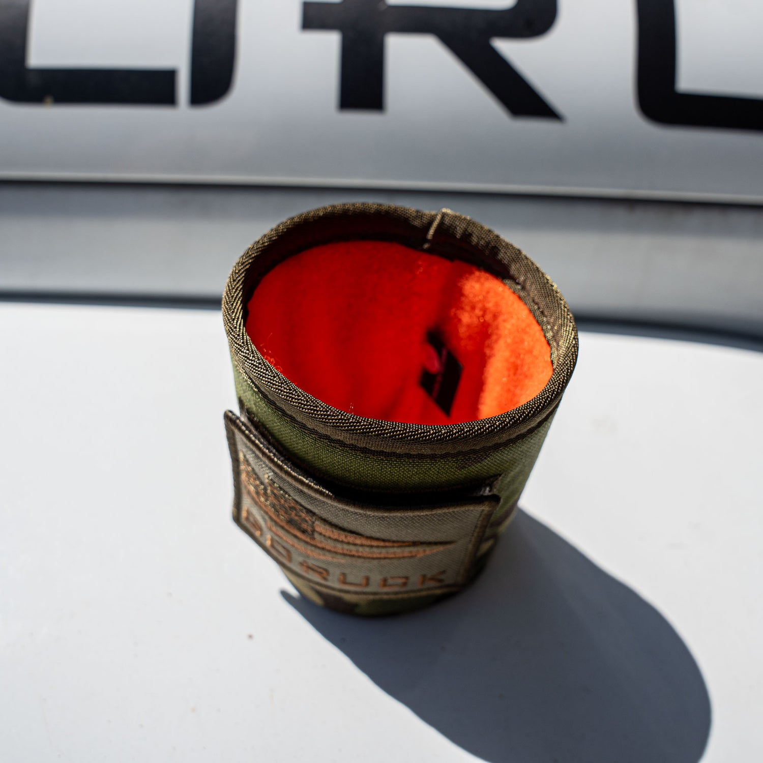 The Ballistic Beer Jacket, a green fabric koozie with an orange lining, rests on a white surface in sunlight.
