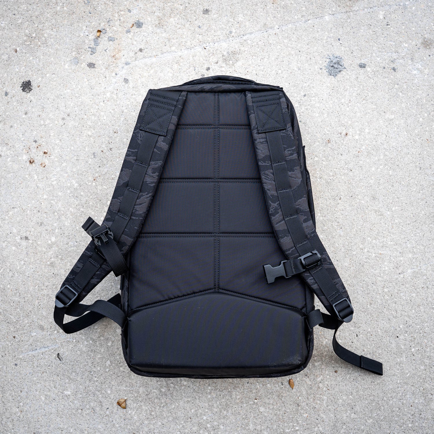 The Rucker 5.0, a black rucksack with padded back, lumbar support, and comfortable shoulder straps, lies face down on light concrete—perfect for rucking or everyday use.