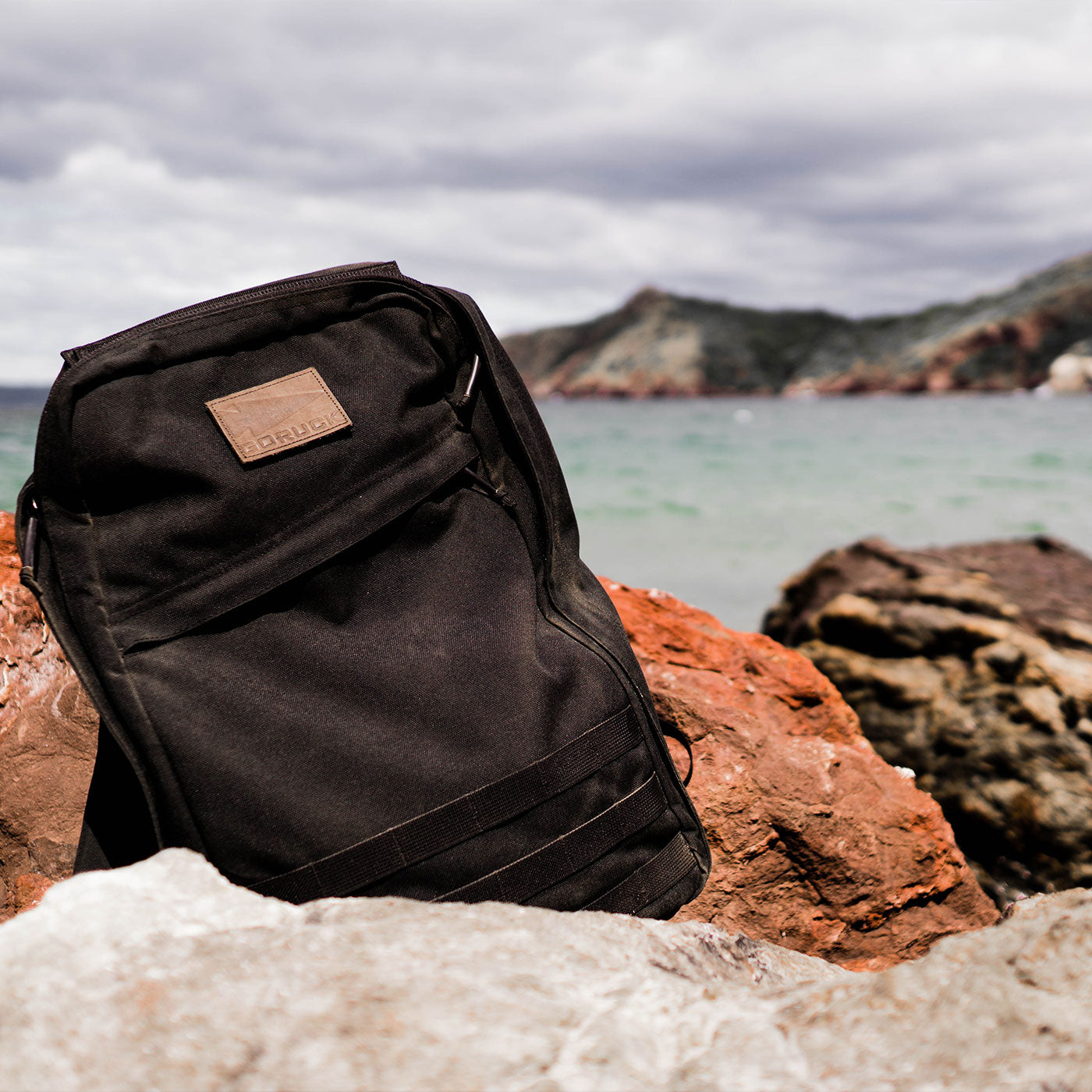 A black backpack rests on reddish rocks by the sea, with mountains and cloudy skies in the background.