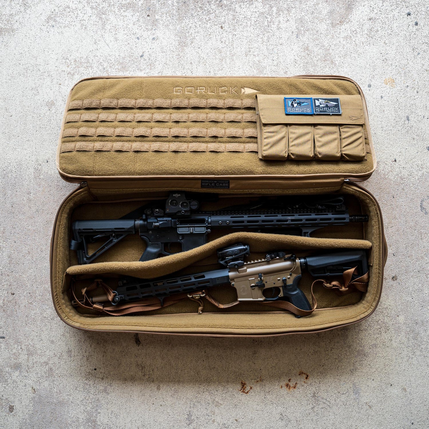 A Rifle Case in tan, adorned with patches, lies open on a concrete floor, displaying two rifles inside.