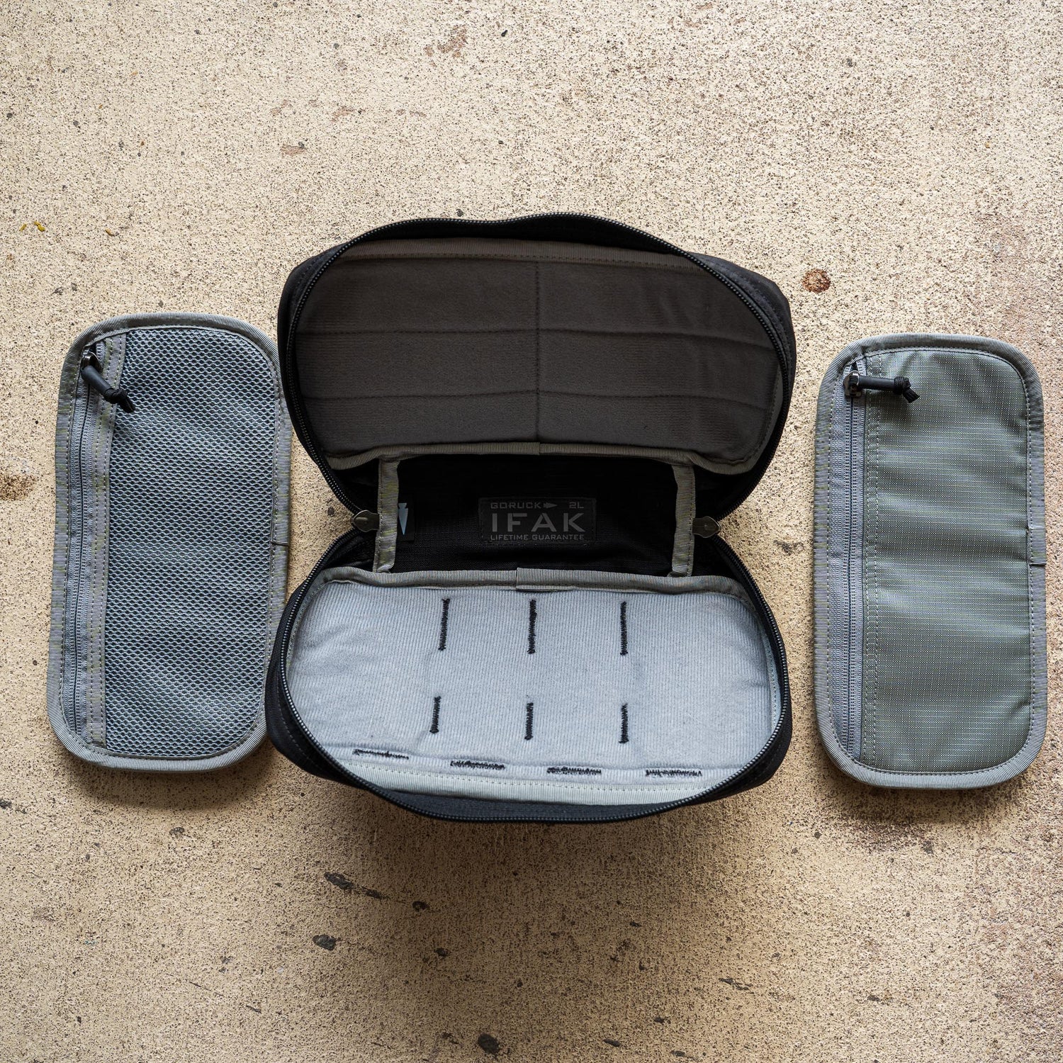 The IFAK (Black + Red Thread + Grey Interior + Red Patch) ROBIC w/ Aquaguard is open to show its gray pouches on each side, displayed on a textured beige surface.