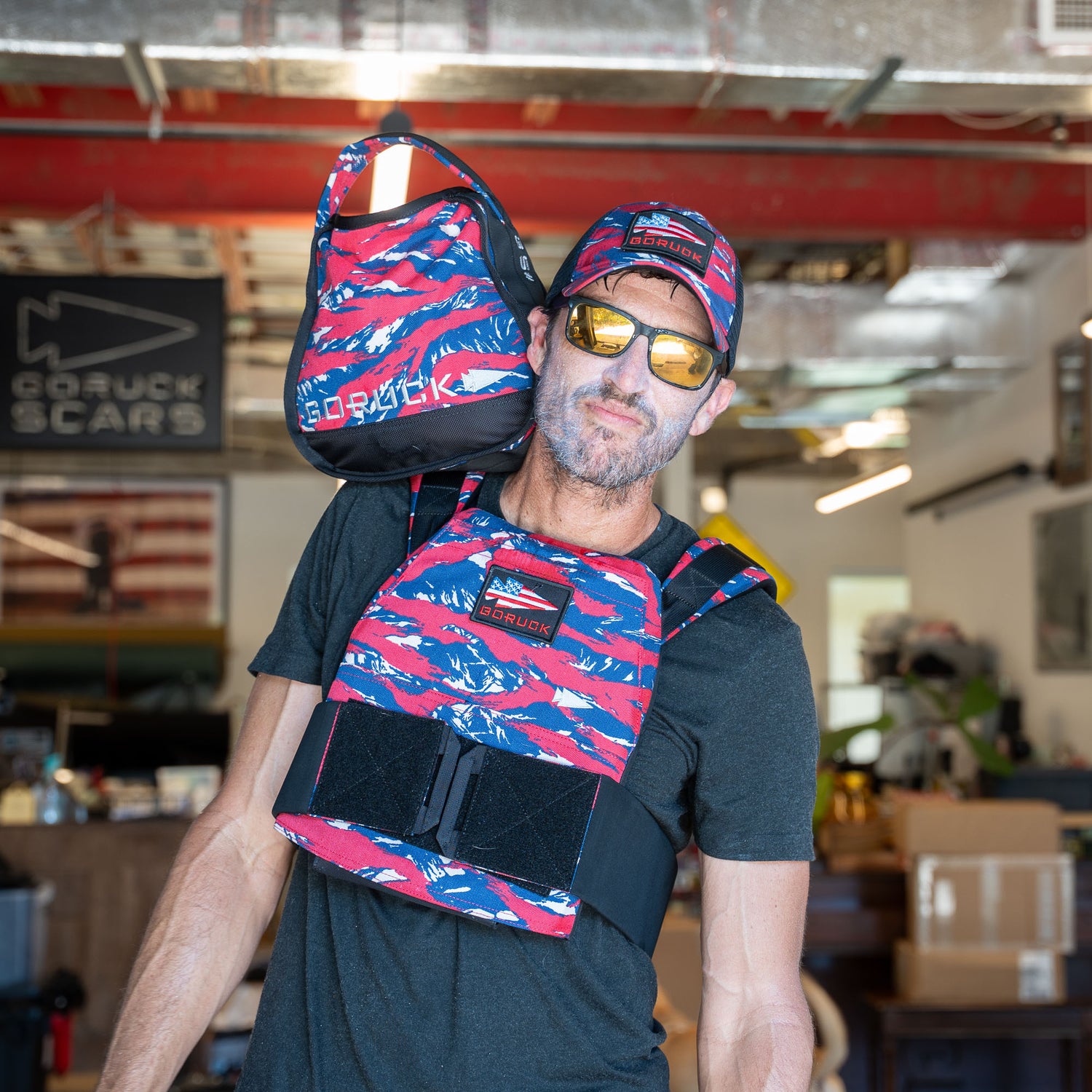 Man carrying a Sand Kettlebells bag with padded handle over his shoulder indoors, wearing sunglasses and red, white, and blue gear.