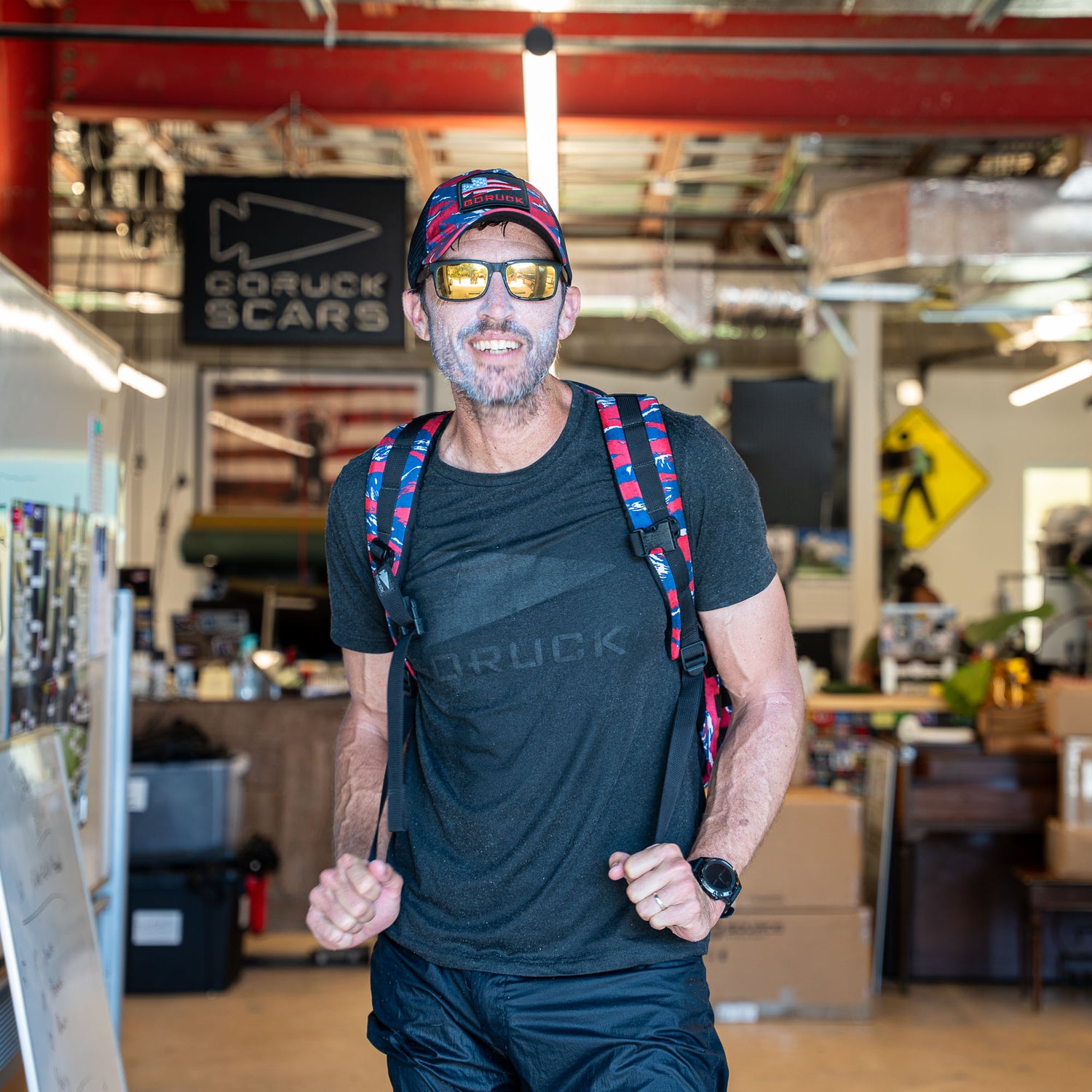 Smiling man in sunglasses and backpack stands indoors at a GORUCK SCARS location, ready for rucking with his Rucker 5.0.
