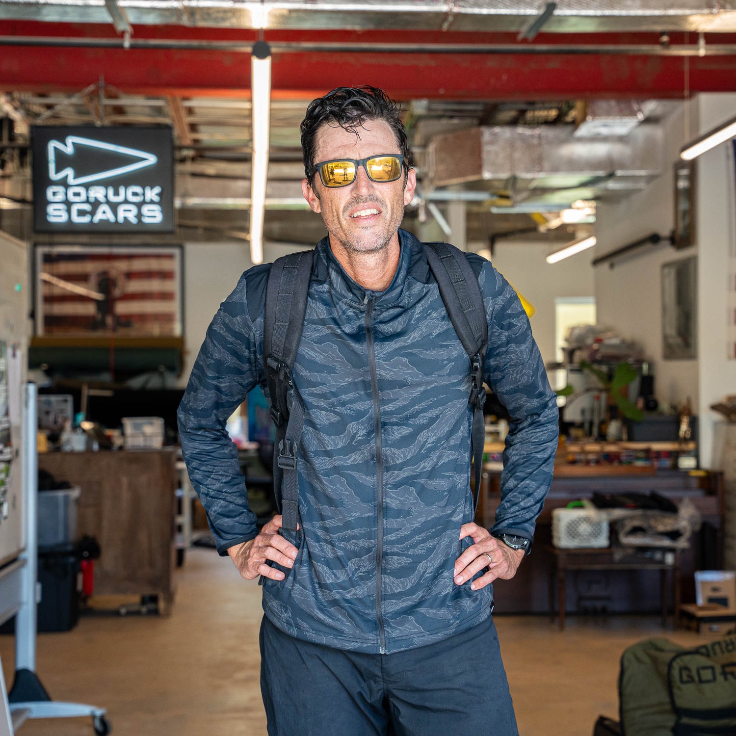 A man wearing sunglasses and a backpack stands in a workshop with a
