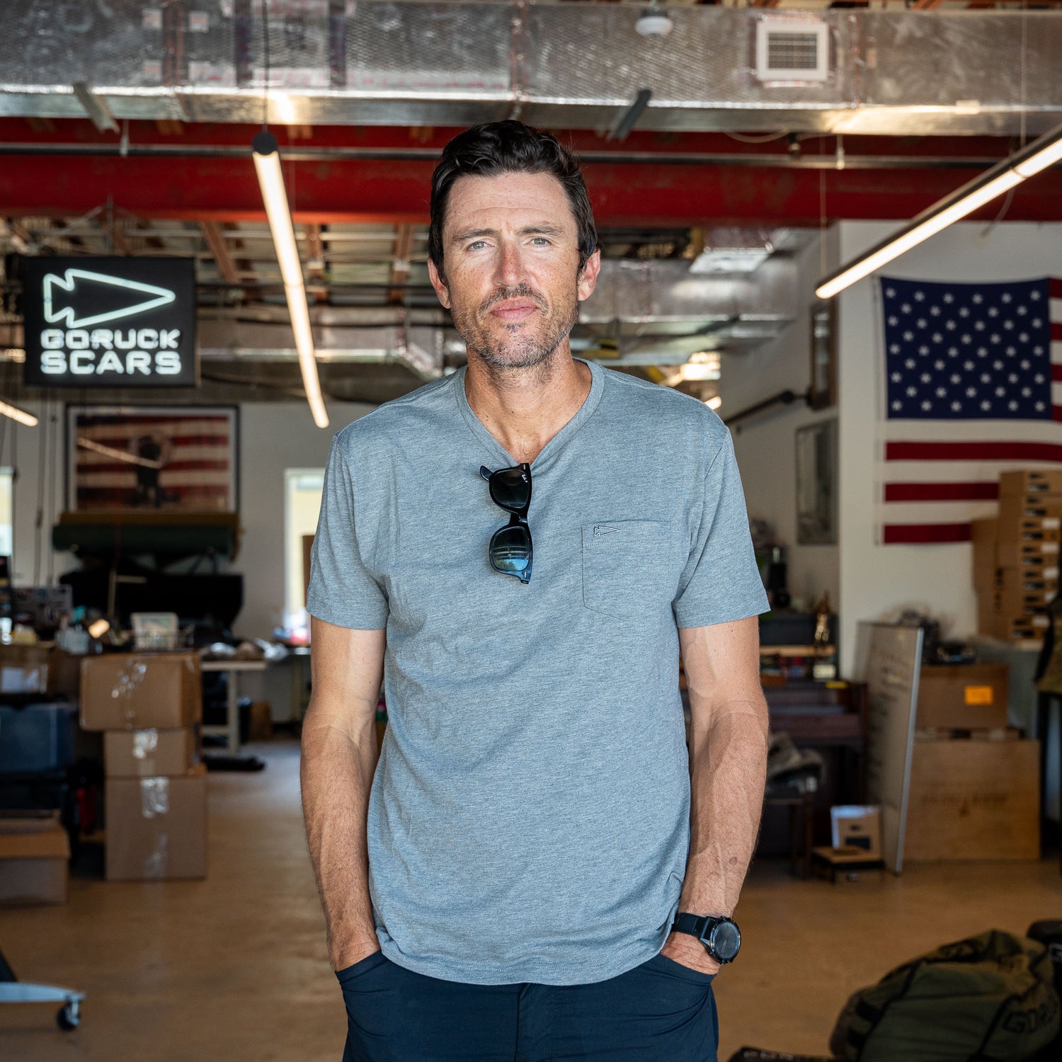 A man wearing the Short Sleeve Pocket Tee - Tri-BlendX stands in a workshop with an American flag and GORUCK SCARS sign in the background, embodying classic simplicity and a versatile spirit.