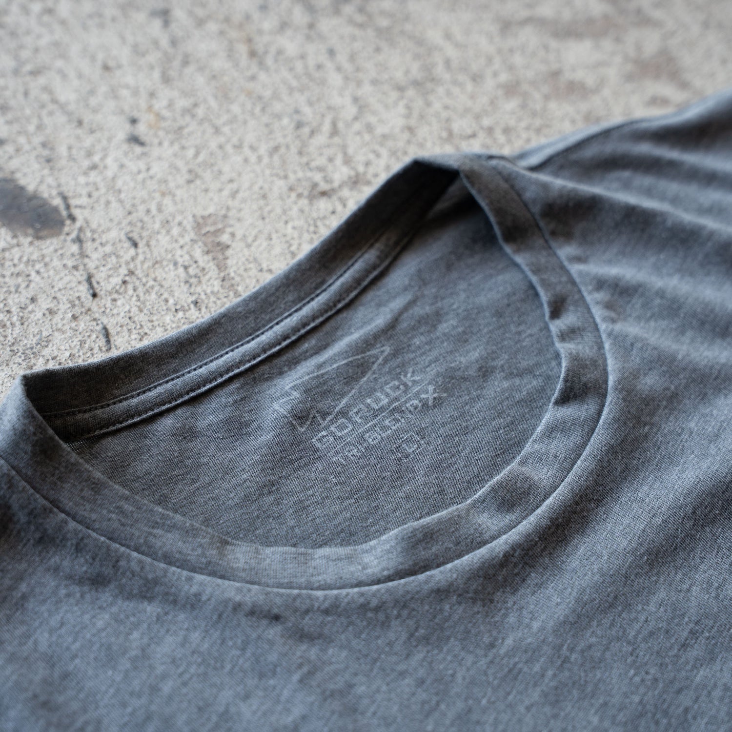 Close-up of a blue-gray Short Sleeve Pocket Tee - Tri-BlendX collar and tag, lying flat on a textured, light-colored surface—showcasing the versatile, classic simplicity that defines Ruck or Die's timeless style.