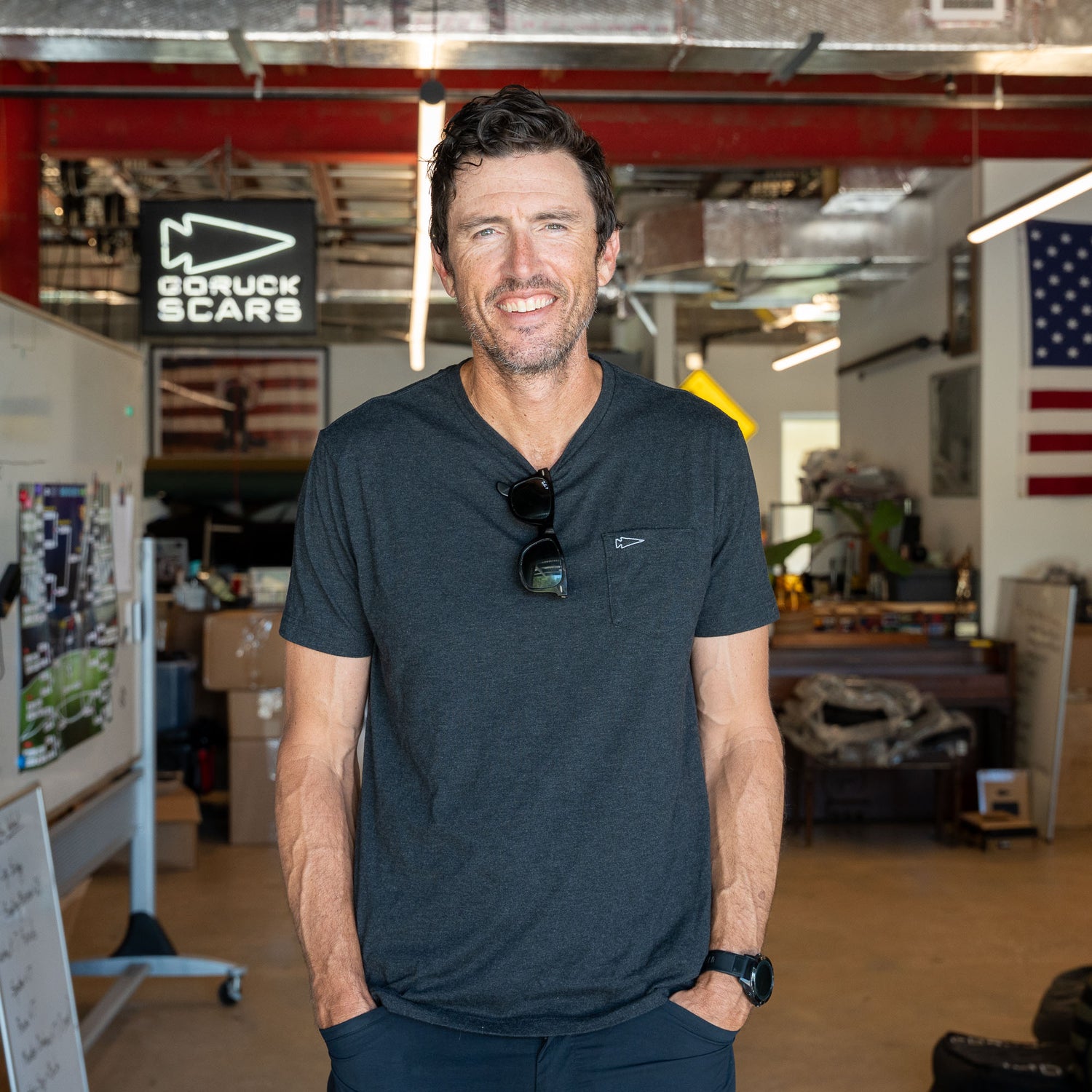 A man stands smiling in a workshop, embodying classic simplicity in the Short Sleeve Pocket Tee - Tri-BlendX, with an American flag and GORUCK SCARS sign behind him.