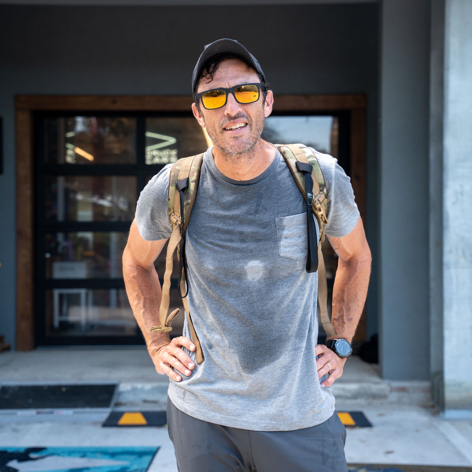 A man wearing sunglasses, a backpack, and the Short Sleeve Pocket Tee - Tri-BlendX in gray stands outside a building, looking at the camera with an air of classic simplicity.