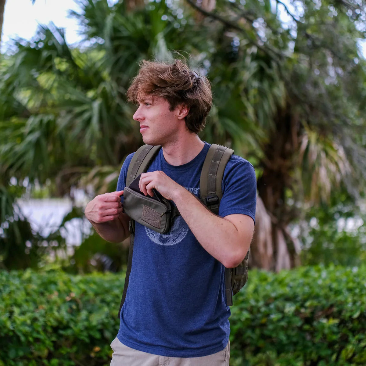 Man wearing GORUCK rucksack outdoors, holding pouch against green leafy background