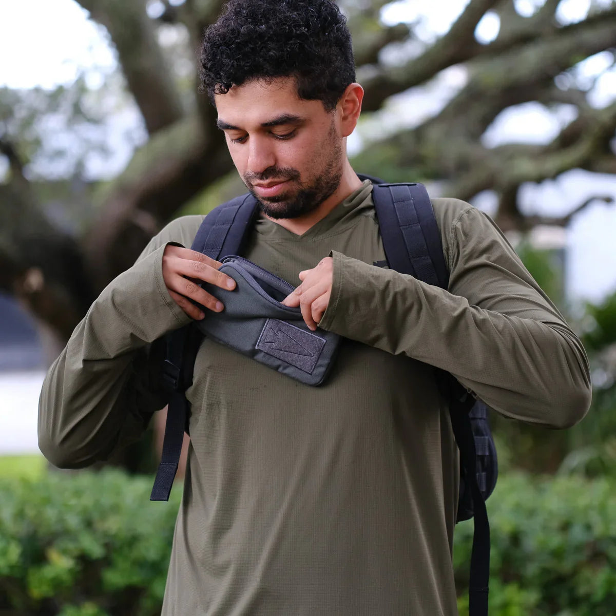 Man wearing GORUCK rucking backpack and khaki shirt, opening gear pouch outdoors