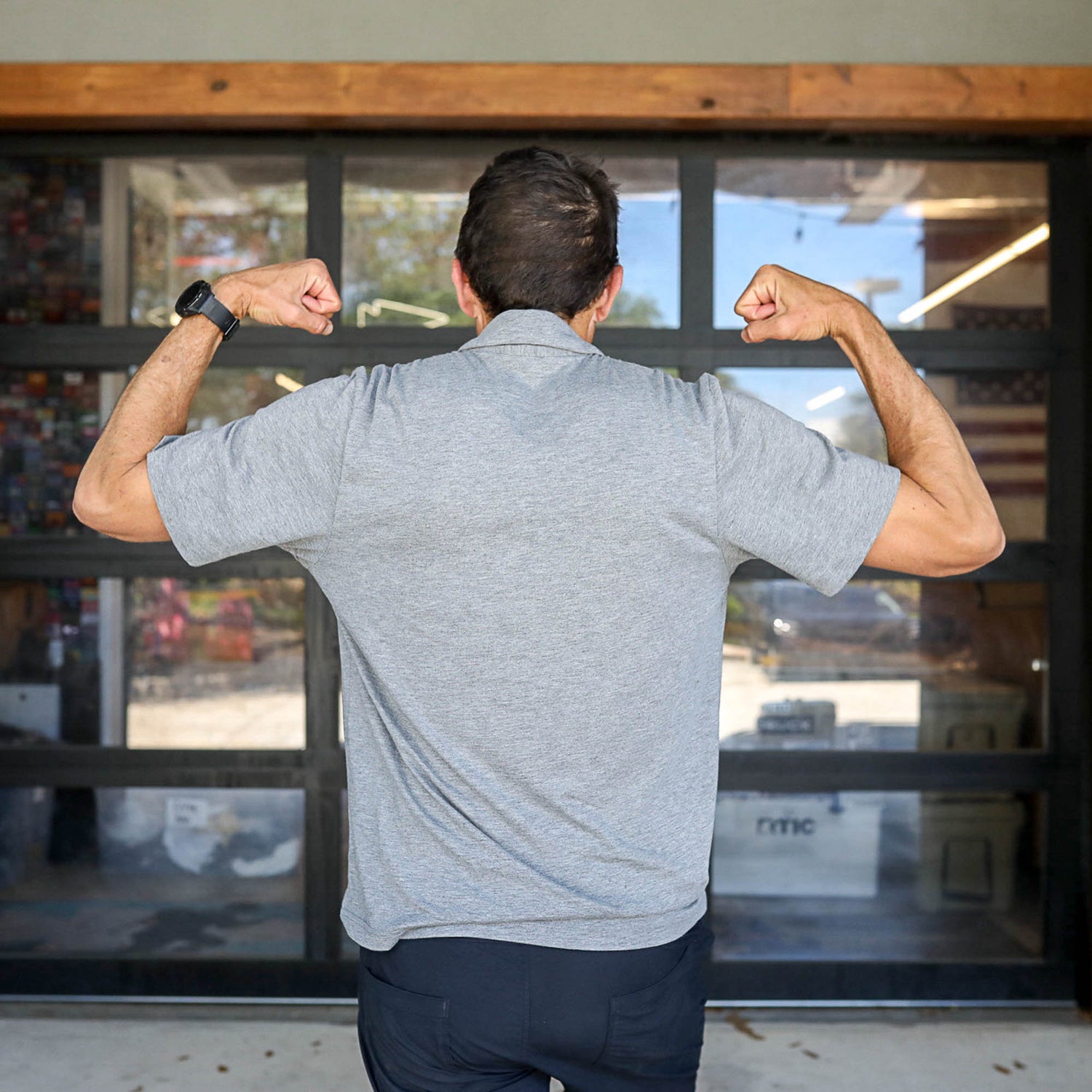 Man wearing a gray Men's Polo with Pocket - Tri-BlendX flexes his arms and shows his back while standing in front of glass doors.