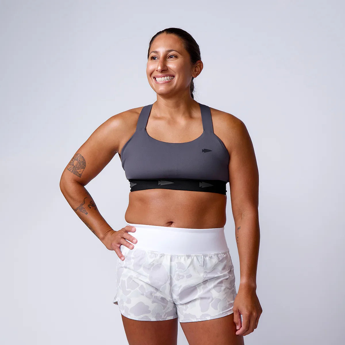 Smiling woman in gray GORUCK sports bra and camo shorts, studio background, rucking gear