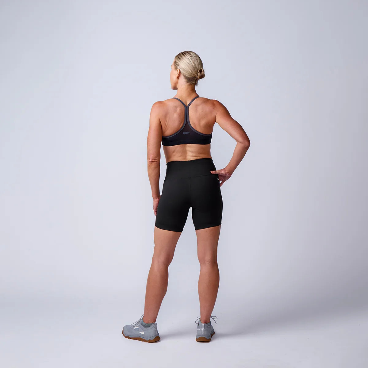 Athletic woman in black GORUCK shorts and sports bra, standing in studio, rear view.