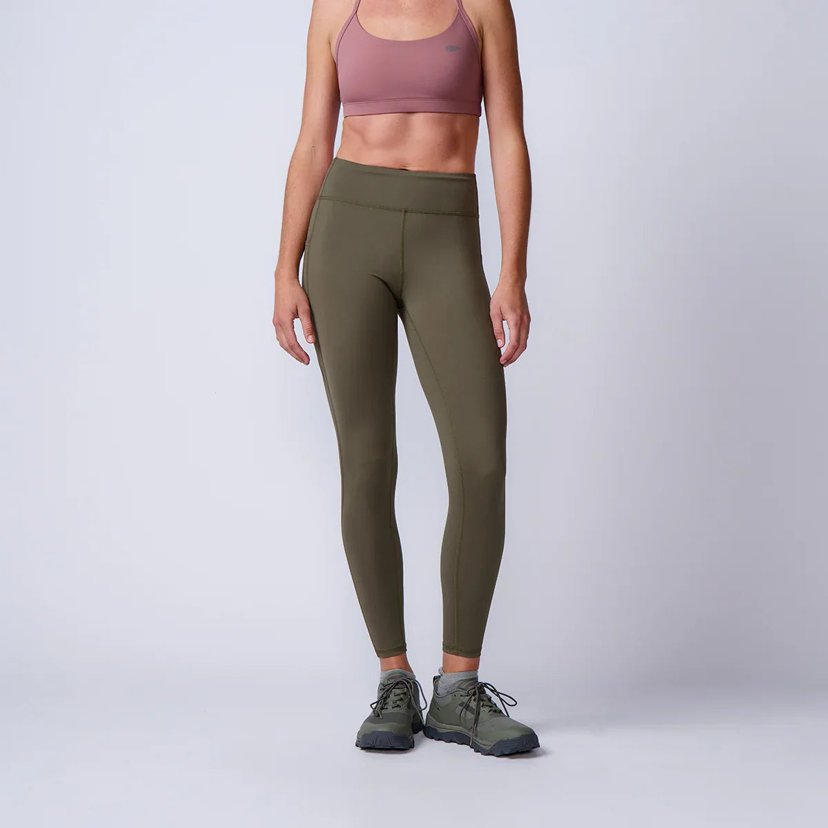 Woman wearing olive green GORUCK leggings, pink sports bra, rucking shoes in studio setting