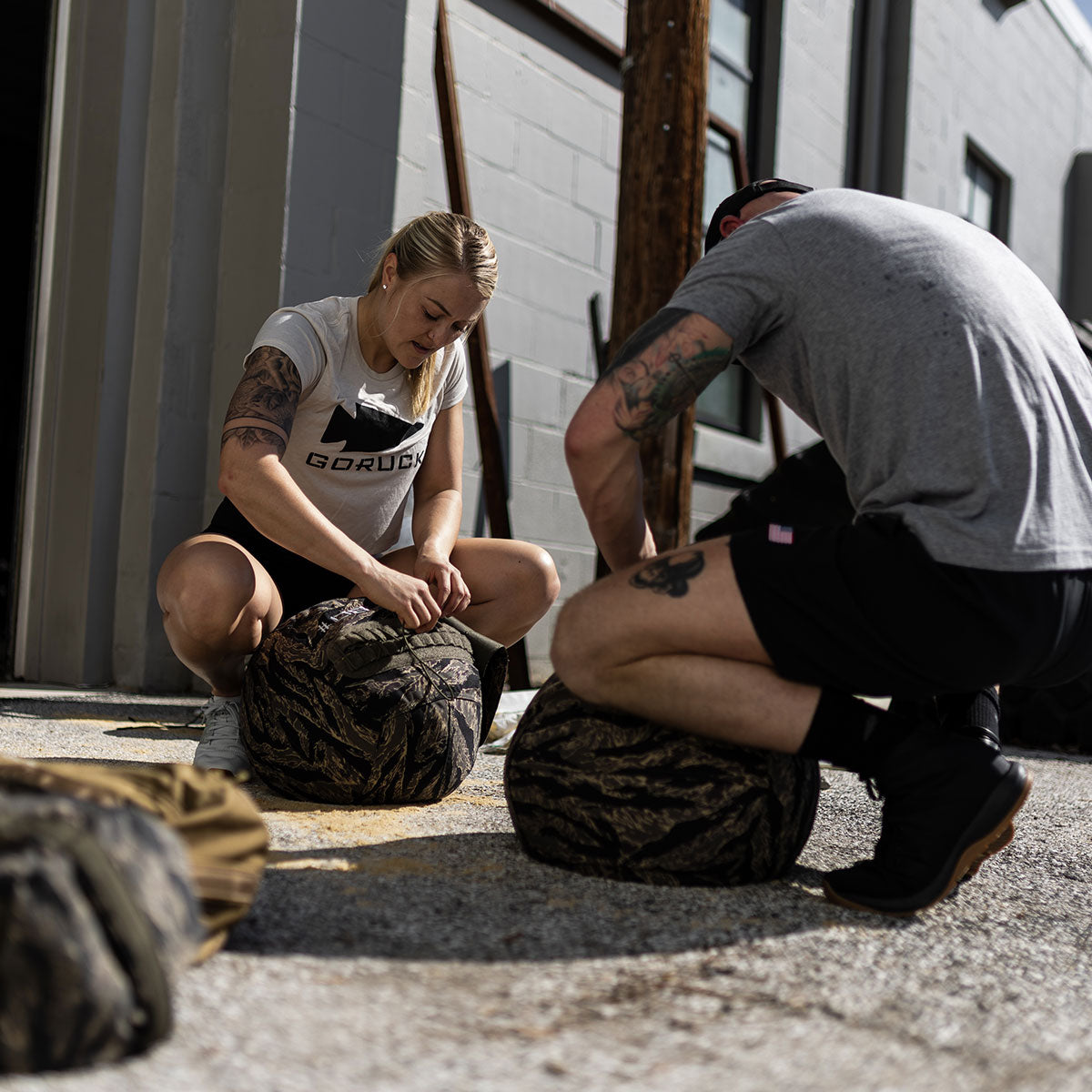 Two people kneel outdoors, packing sandbags on a sunny day beside a gray building.