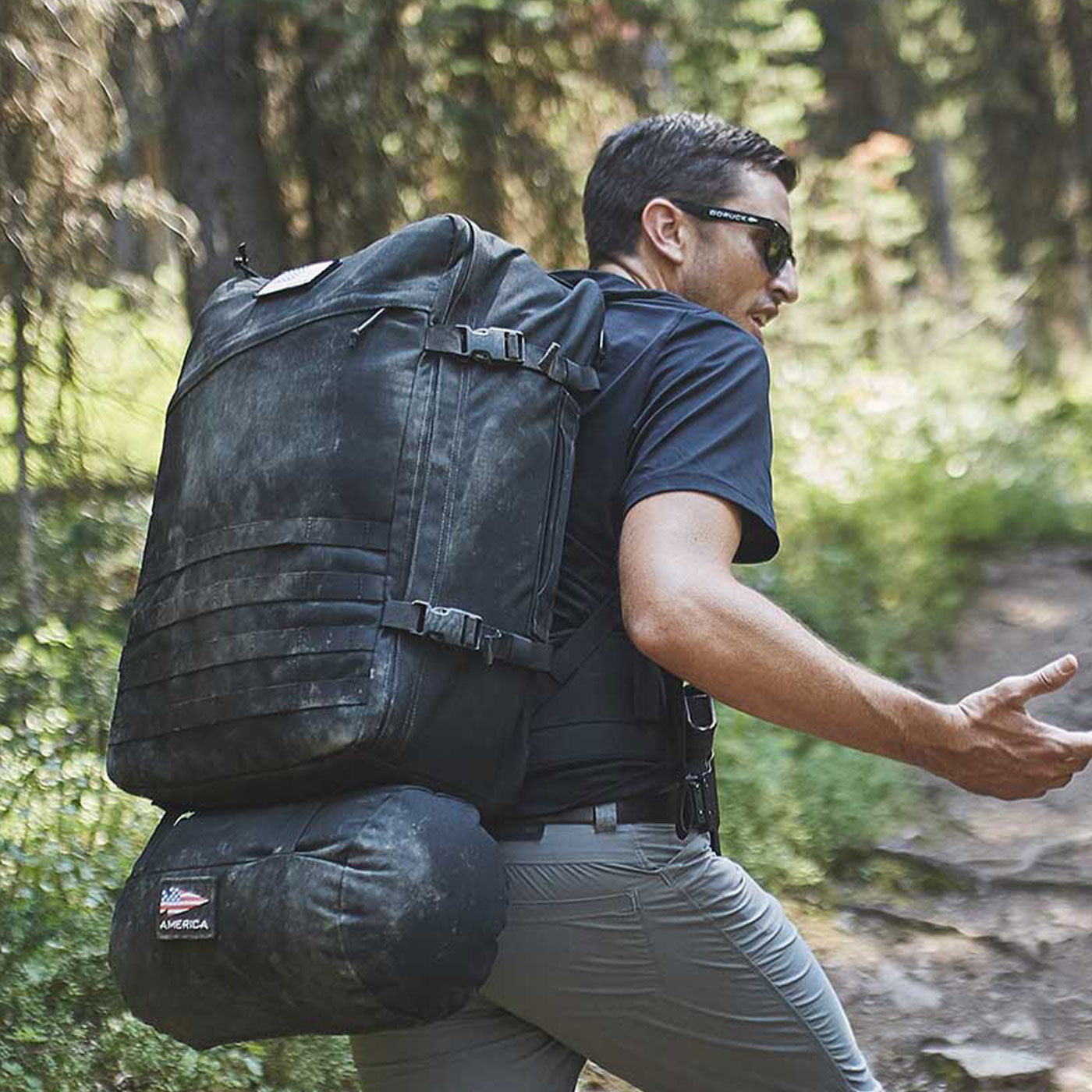 Man wearing sunglasses and a large hiking backpack walks on a forest trail in sunlight.