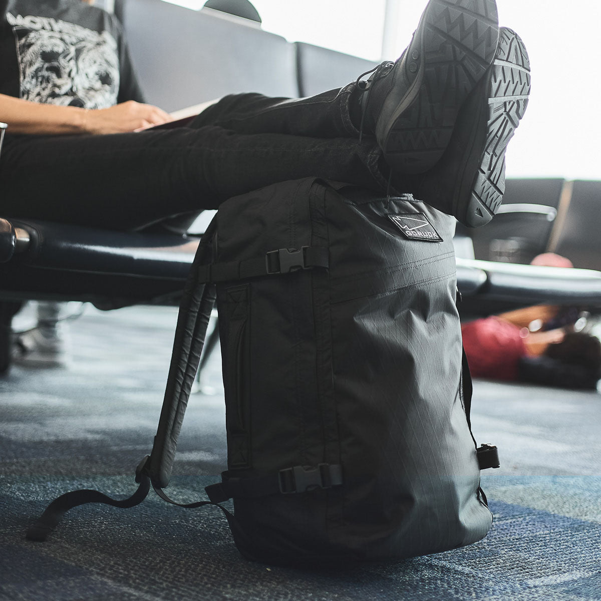 Person resting feet on a black backpack at an airport, sitting on a bench, with luggage in the background.