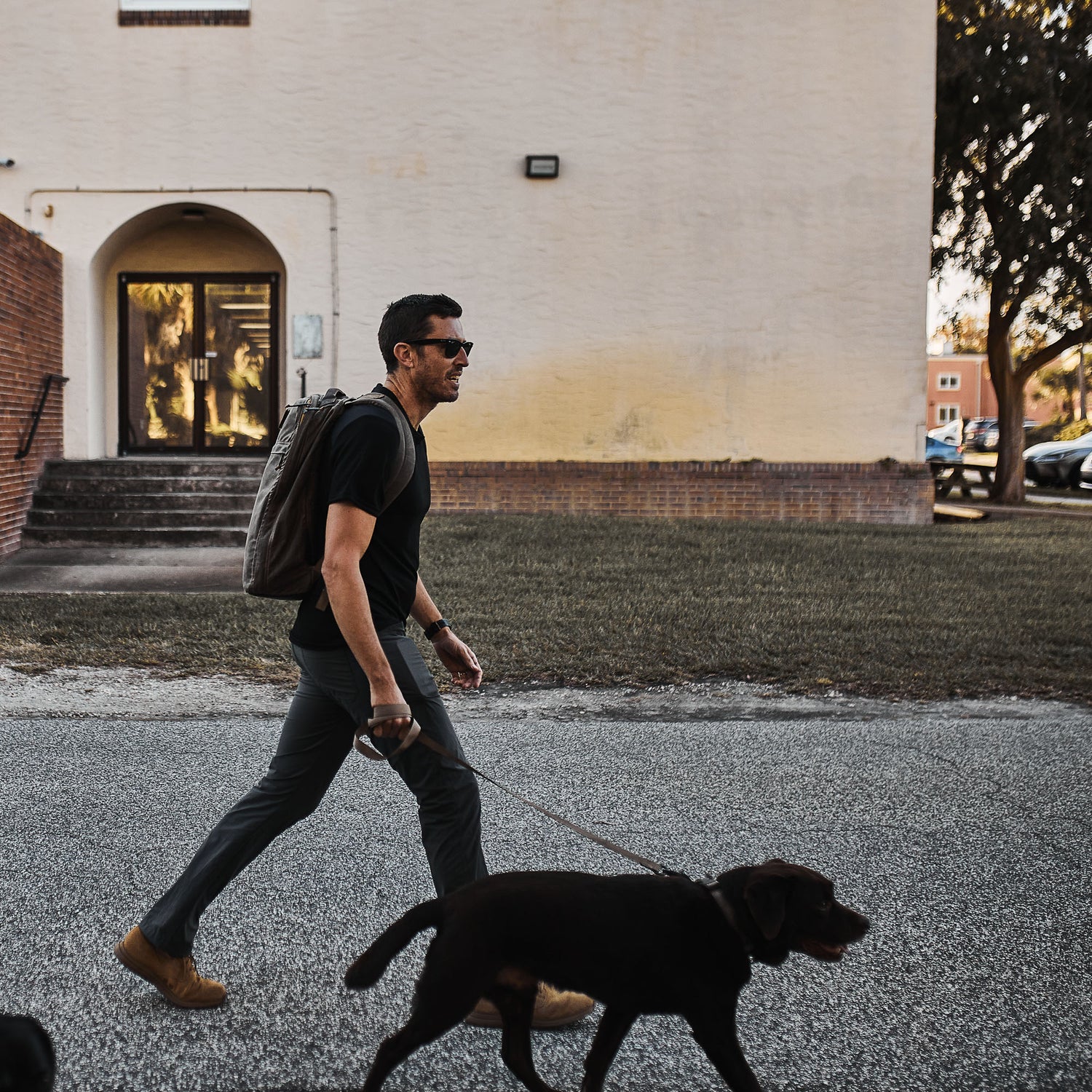 A man in sunglasses wearing the Men’s Commando Tee - Merino Wool with reinforced shoulders, carrying a backpack and walking a black dog on a leash along a street near a building.