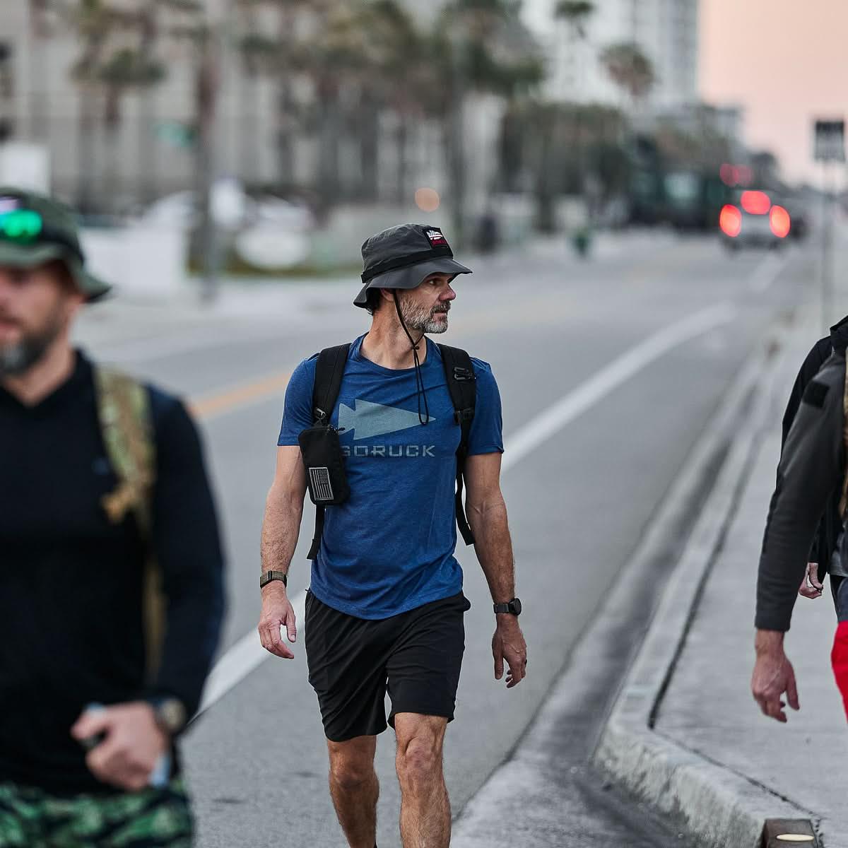 Man wearing charcoal GORUCK boonie hat and blue GORUCK shirt walking outdoors on city street