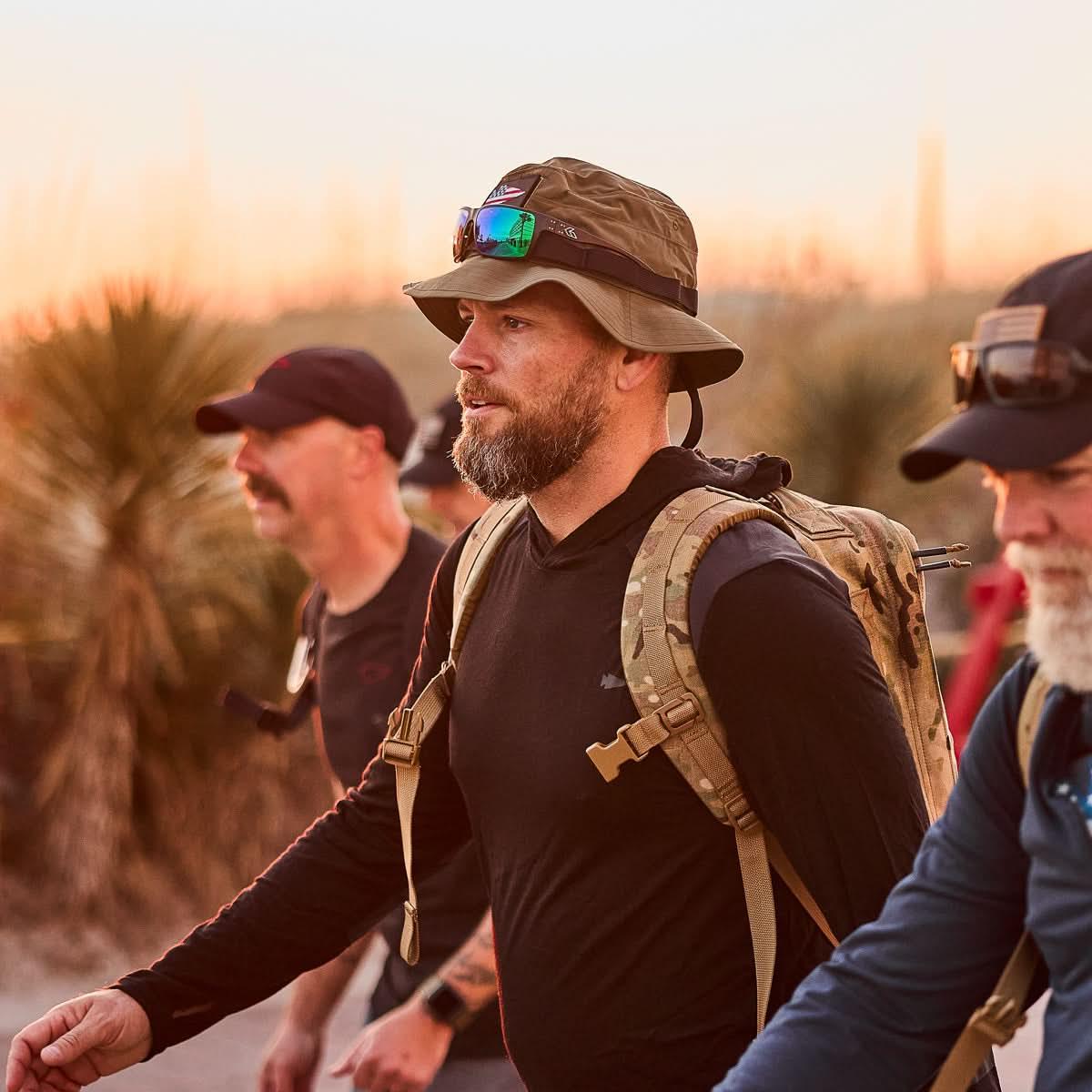 Man wearing ranger green boonie hat, black shirt, and camouflage backpack, rucking outdoors at sunset, GORUCK gear in use.
