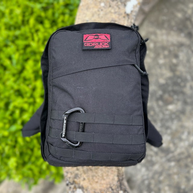 The GR2 backpack in black, featuring a carabiner, sits on stone near green foliage—ideal for one bag travel and is fully carry-on compliant.