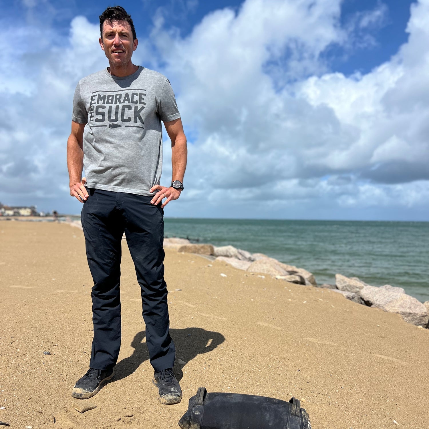 A man stands on a sandy beach near rocks and the ocean, wearing a shirt that says