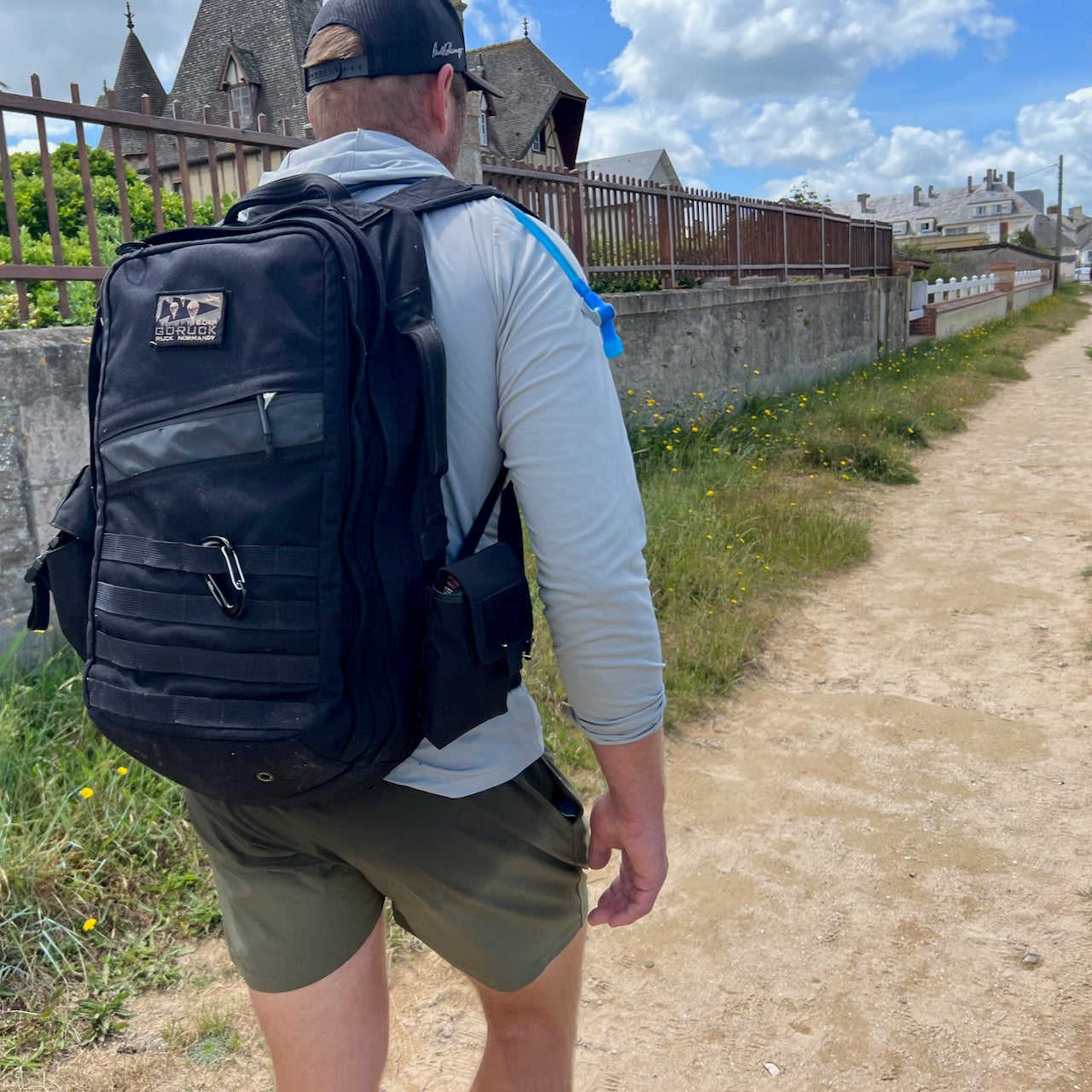 A man wearing a cap and backpack with a Simple Side Pocket walks on a dirt path near stone buildings and greenery under a partly cloudy sky.