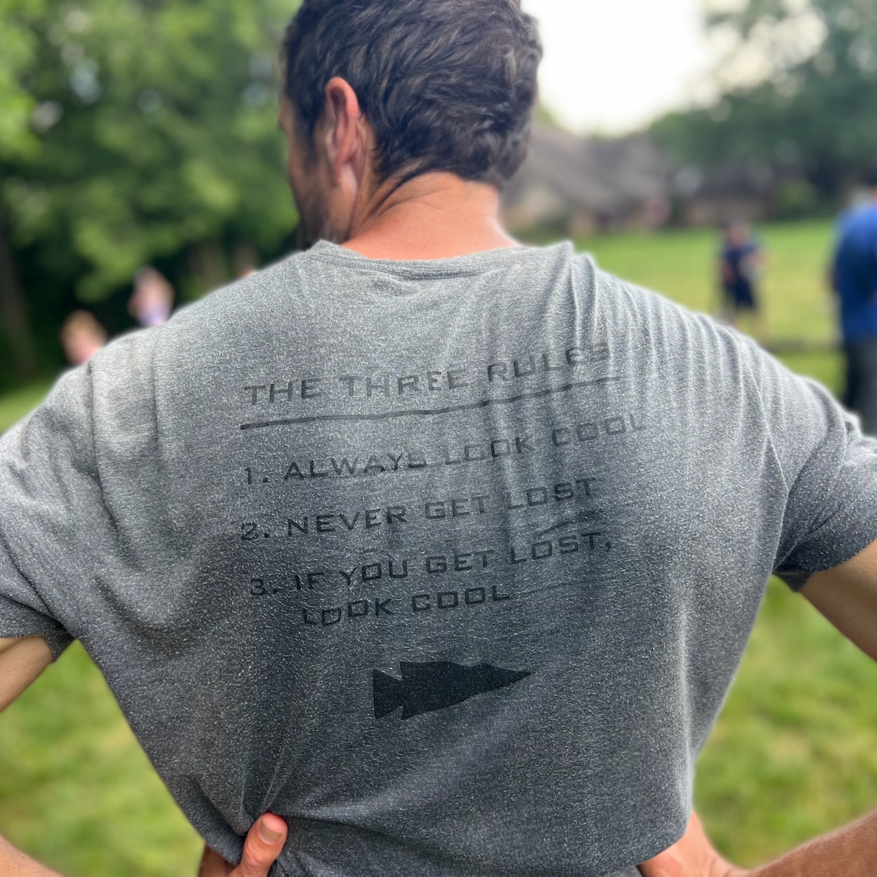 A man stands outdoors wearing the Three Rules Tee - Unisex Tri-BlendX, featuring three rules on the back like