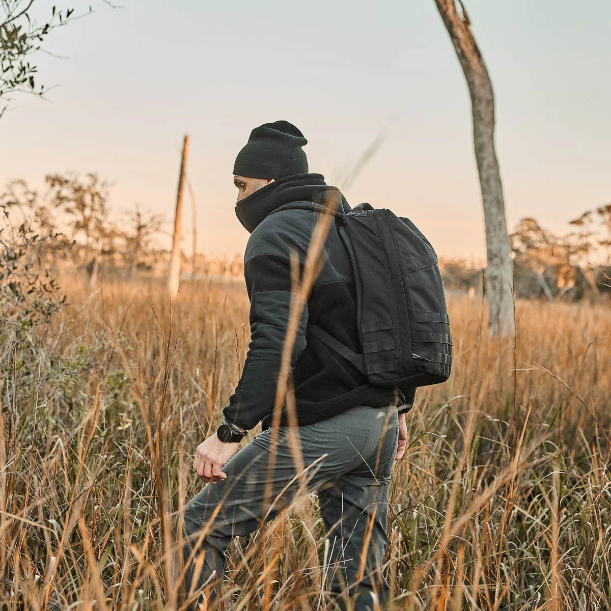 Man rucking outdoors in field at sunset wearing GORUCK backpack, beanie, and jacket