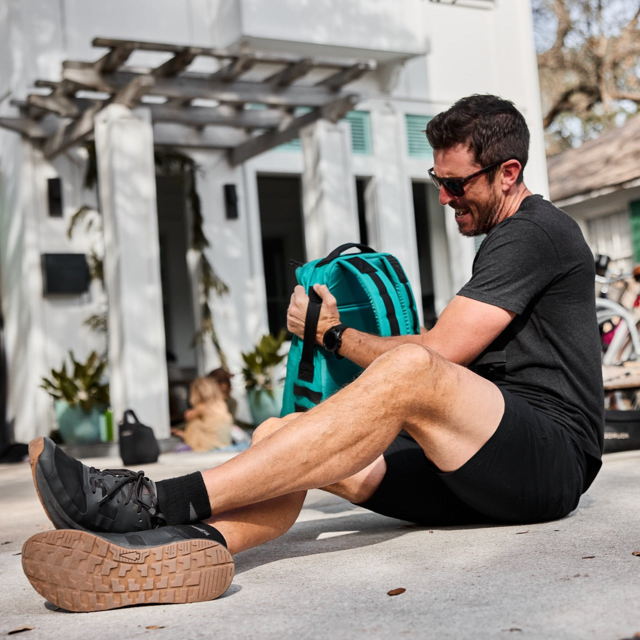 Man in black shirt and shorts laughs while sitting on pavement, holding a teal bag with a house behind—wearing Merino Challenge Socks - Trail Cuff for ultimate comfort on the go.