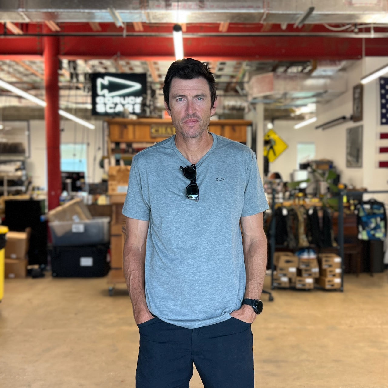 A man wearing the Subtle Spearhead Tee - Unisex Tri-Blend stands indoors in a store or workshop with shelves and boxes in the background, giving off a premium, Special Forces-inspired vibe.