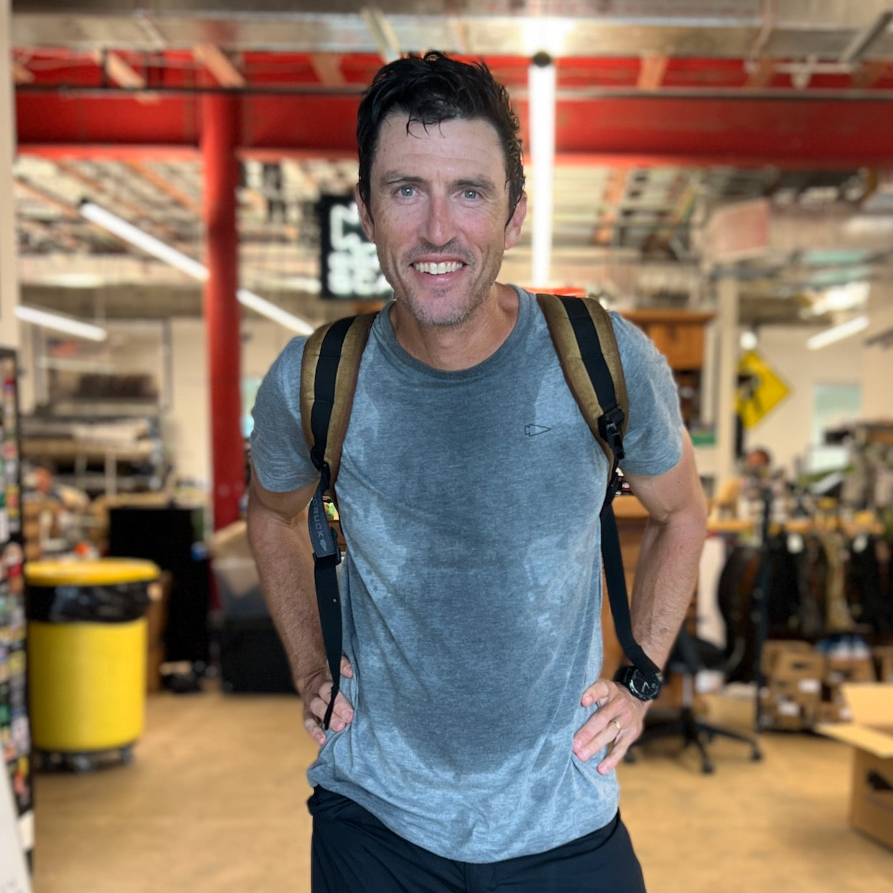 Man smiling in a warehouse, wearing a grey Subtle Spearhead Tee - Unisex Tri-Blend with sweat stains and a backpack, hands on hips.