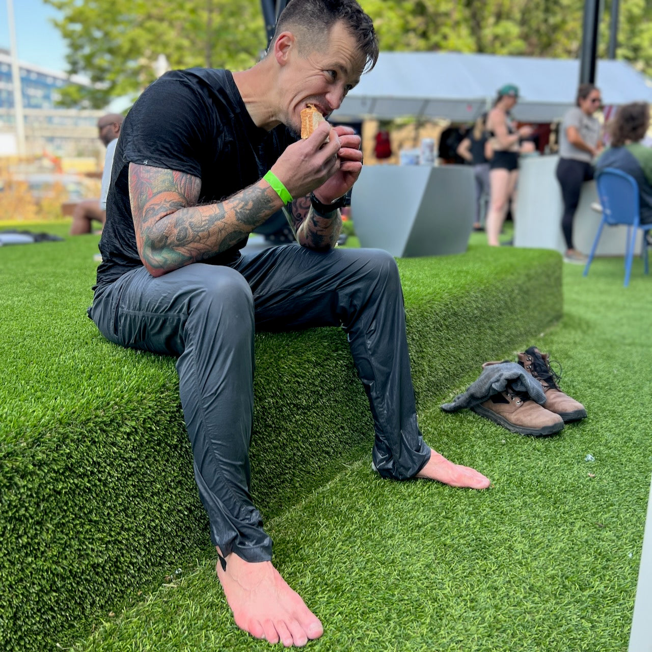 A man in wet clothes sits barefoot on artificial grass, eating a sandwich with his MACV-2 - Mid Top - Chestnut + Black boots and gloves beside him, the aggressive outsole of his Special Forces boots visible.