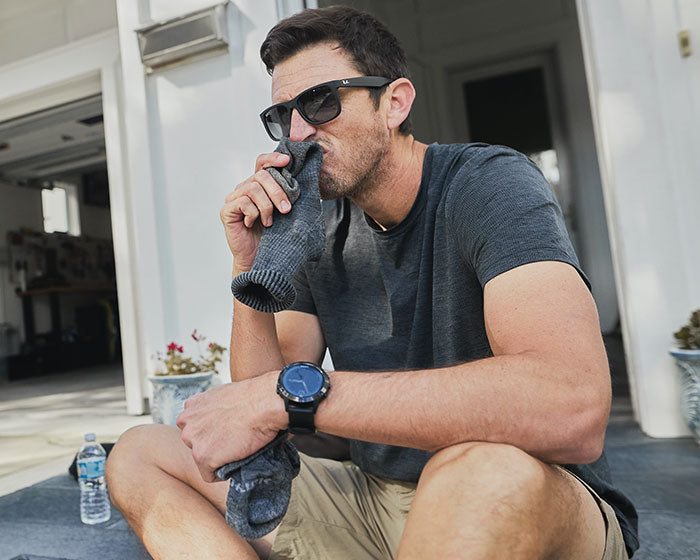 Man wearing sunglasses and a watch sits outside, smelling a gray sock; water bottle beside him.