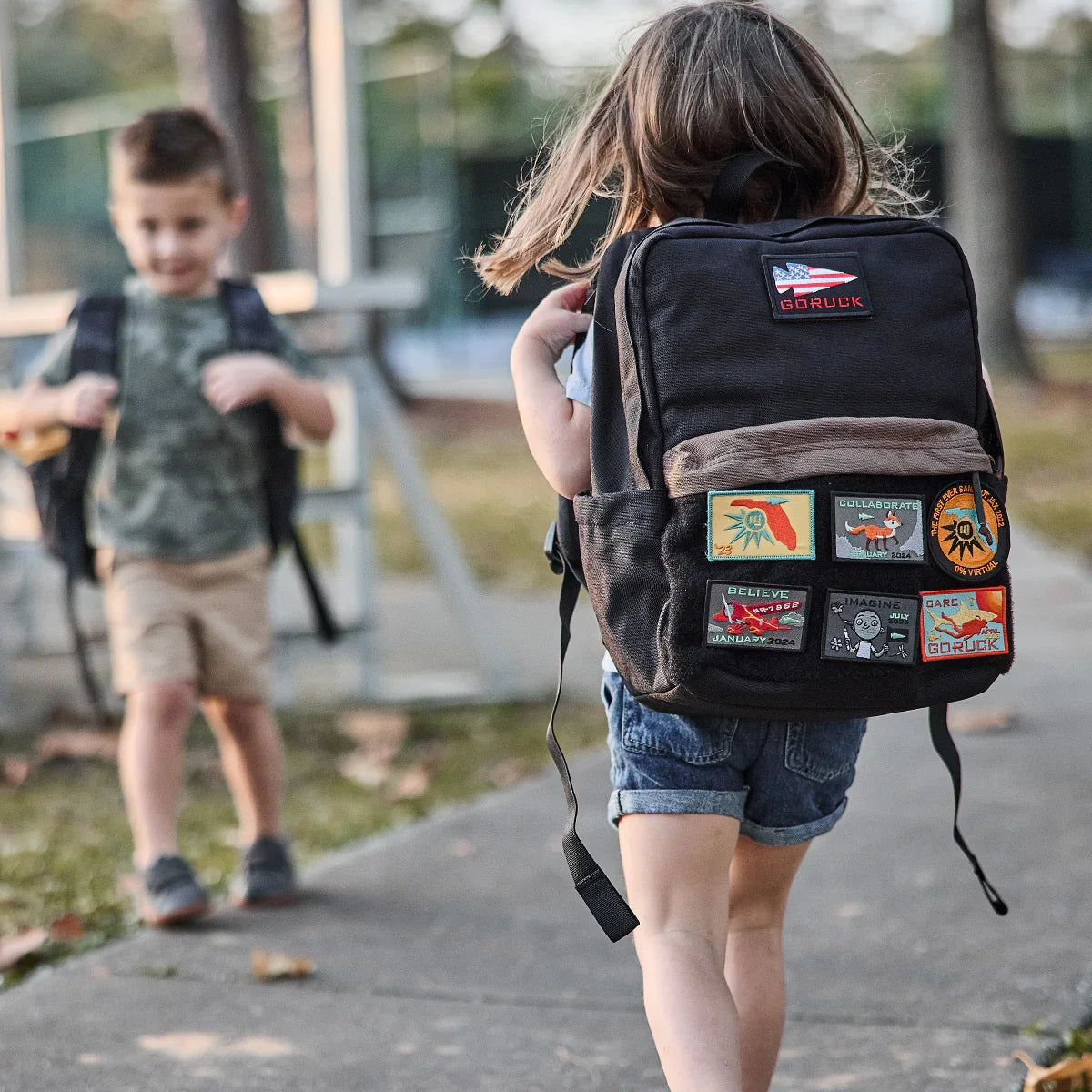 Child wearing black GORUCK backpack with colorful patches, outdoors on a sidewalk