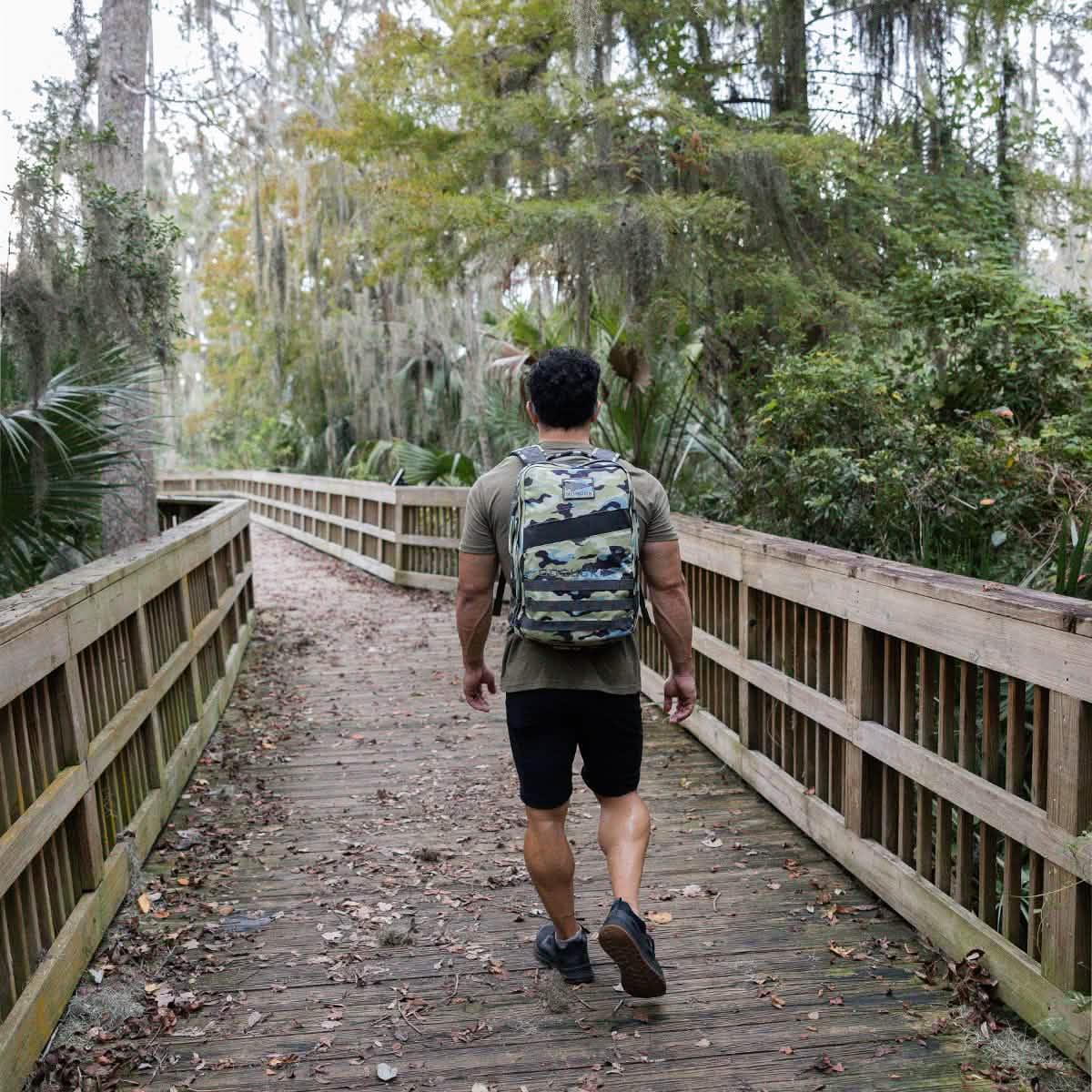 Man rucking in forest with GORUCK camo backpack on wooden trail bridge