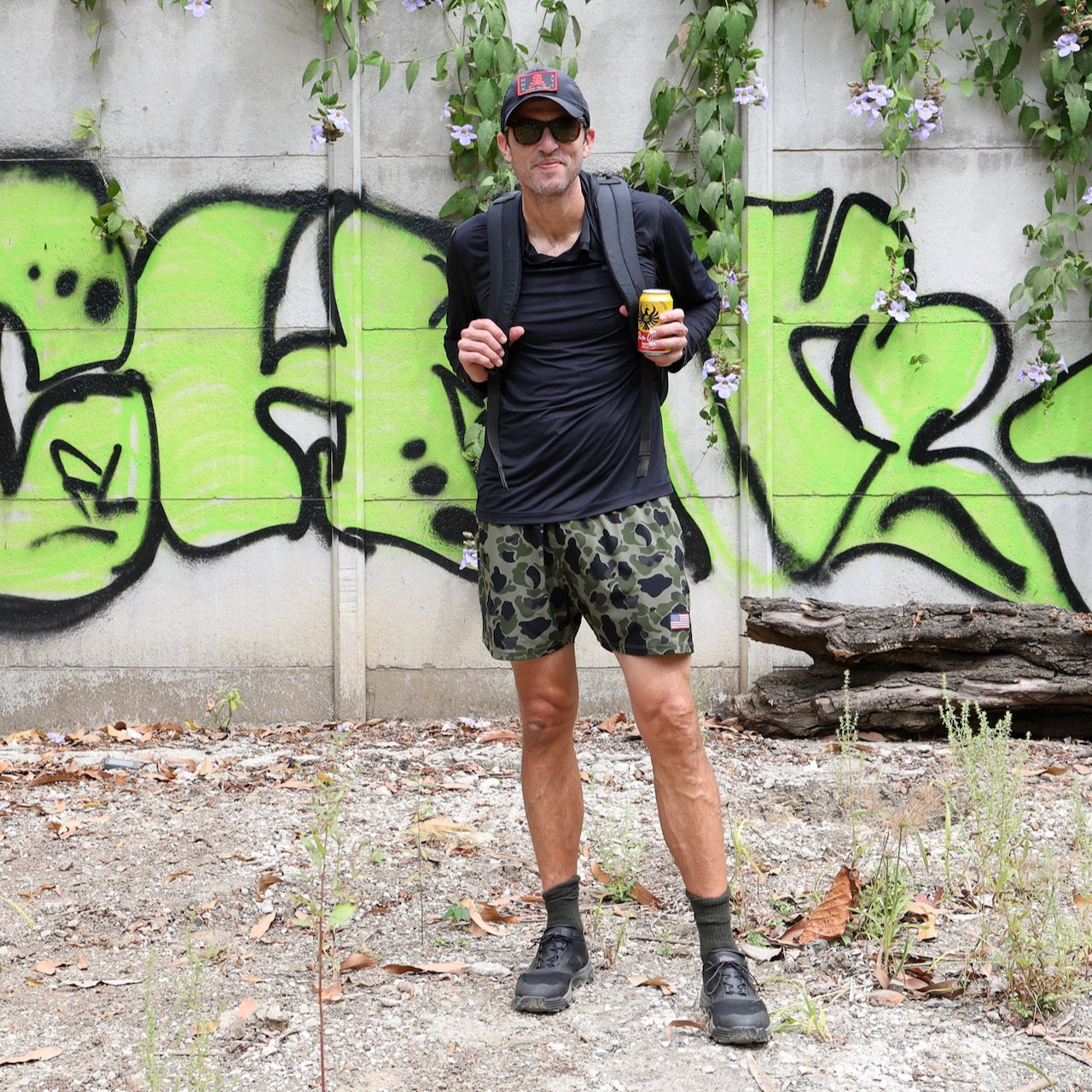 A man in a black shirt, camo shorts, boots, and a backpack stands by a graffiti wall with vines, wearing Mackall - Black + Forged Iron all-terrain rucking shoes.