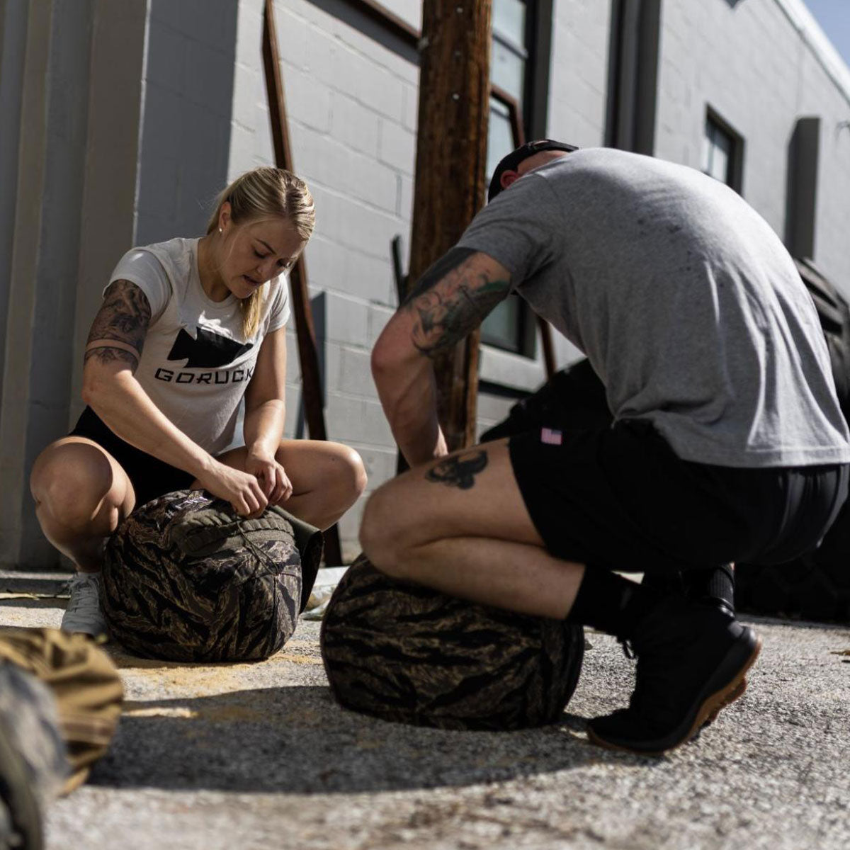 Two people kneeling outdoors, preparing camouflage-patterned gear bags on a sunlit pavement.