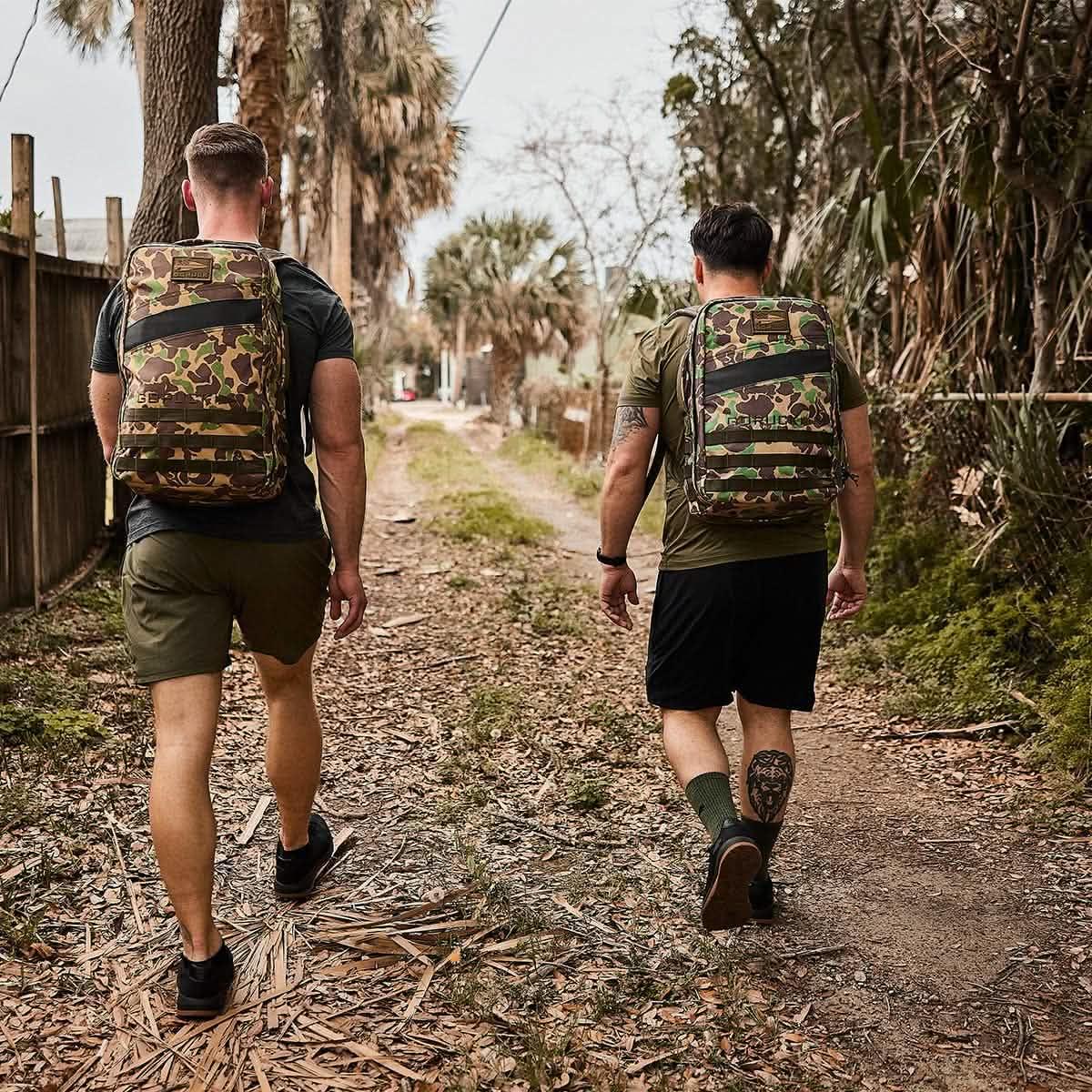 Two men rucking outdoors with GORUCK camo backpacks on a dirt trail, wearing activewear.
