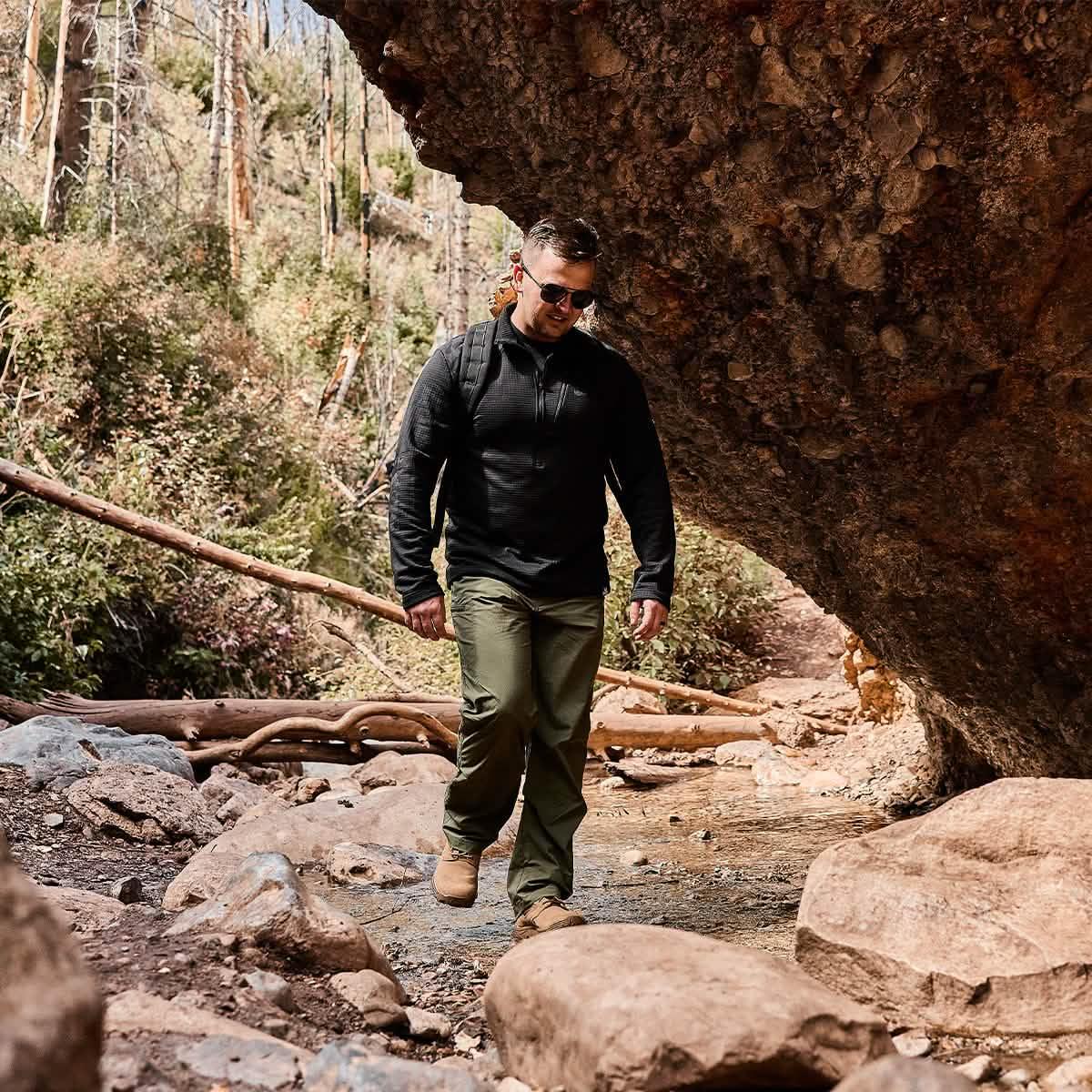 Man hiking outdoors in rugged gear, wearing olive green pants, black jacket, and backpack