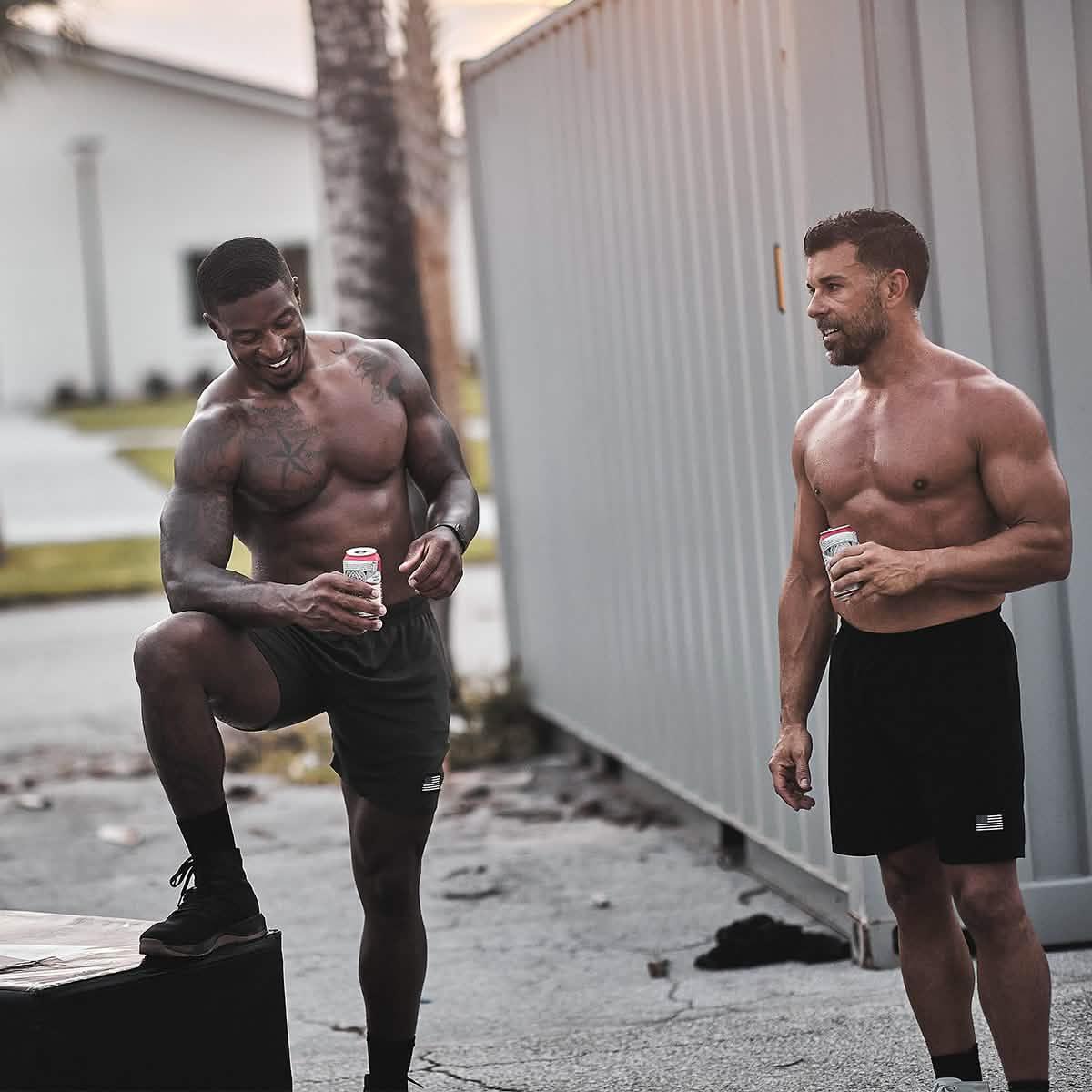 Two muscular men in GORUCK shorts holding drinks outdoors after a workout, smiling and relaxed.